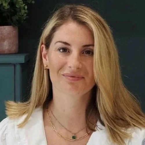 A woman with long blonde hair wearing a white top and layered necklaces, smiling slightly at the camera, sitting in a room with dark teal walls and a plant in the background.