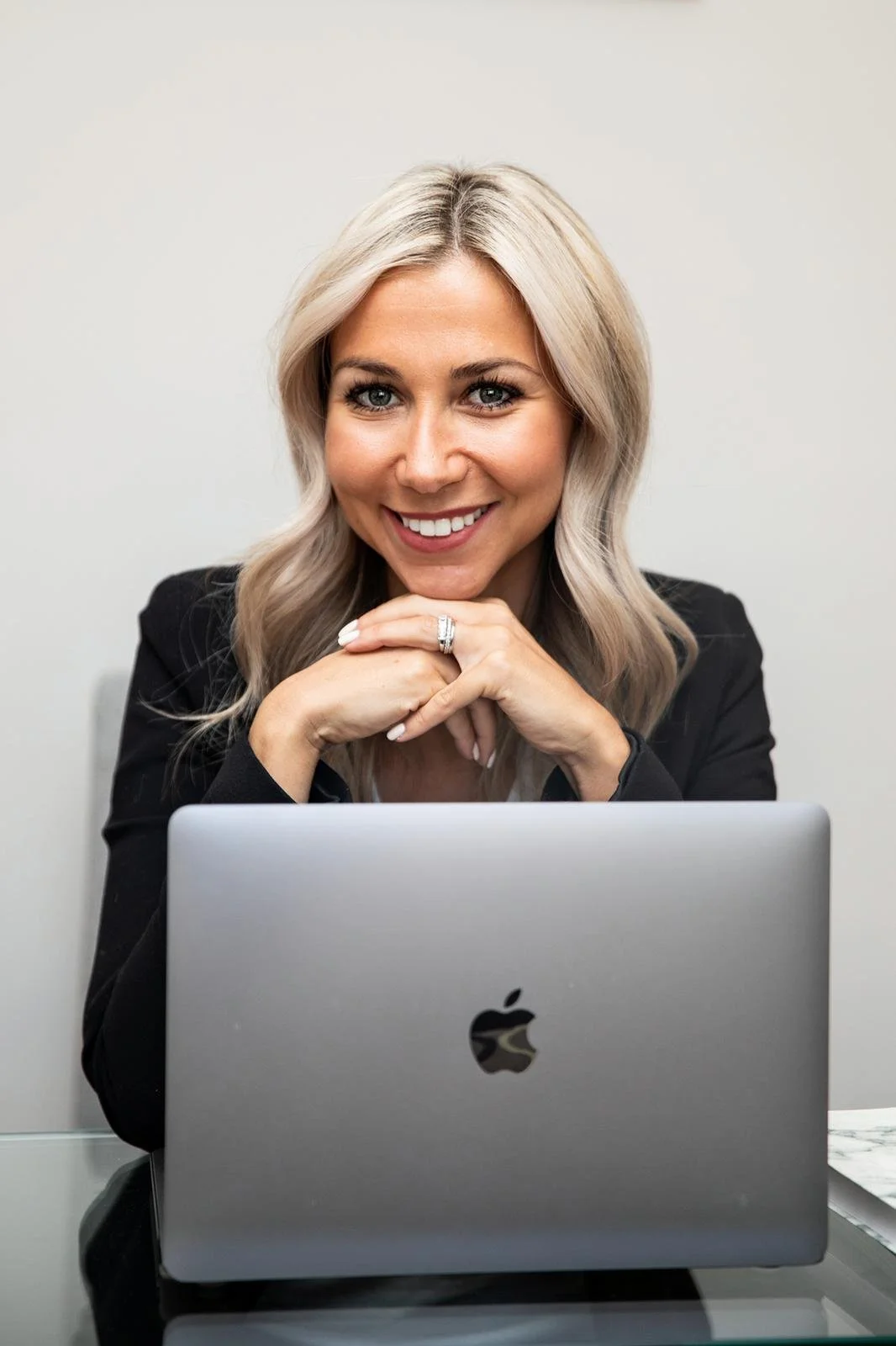 A woman with blonde hair smiling at a laptop, resting her chin on her hands.