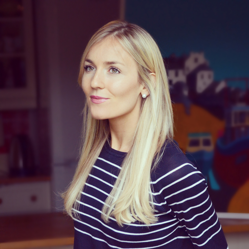 A young woman with long blonde hair, wearing a navy blue and white striped shirt, looking slightly to her left with a gentle smile, in an indoor setting with a blurred background of a colorful painted mural.