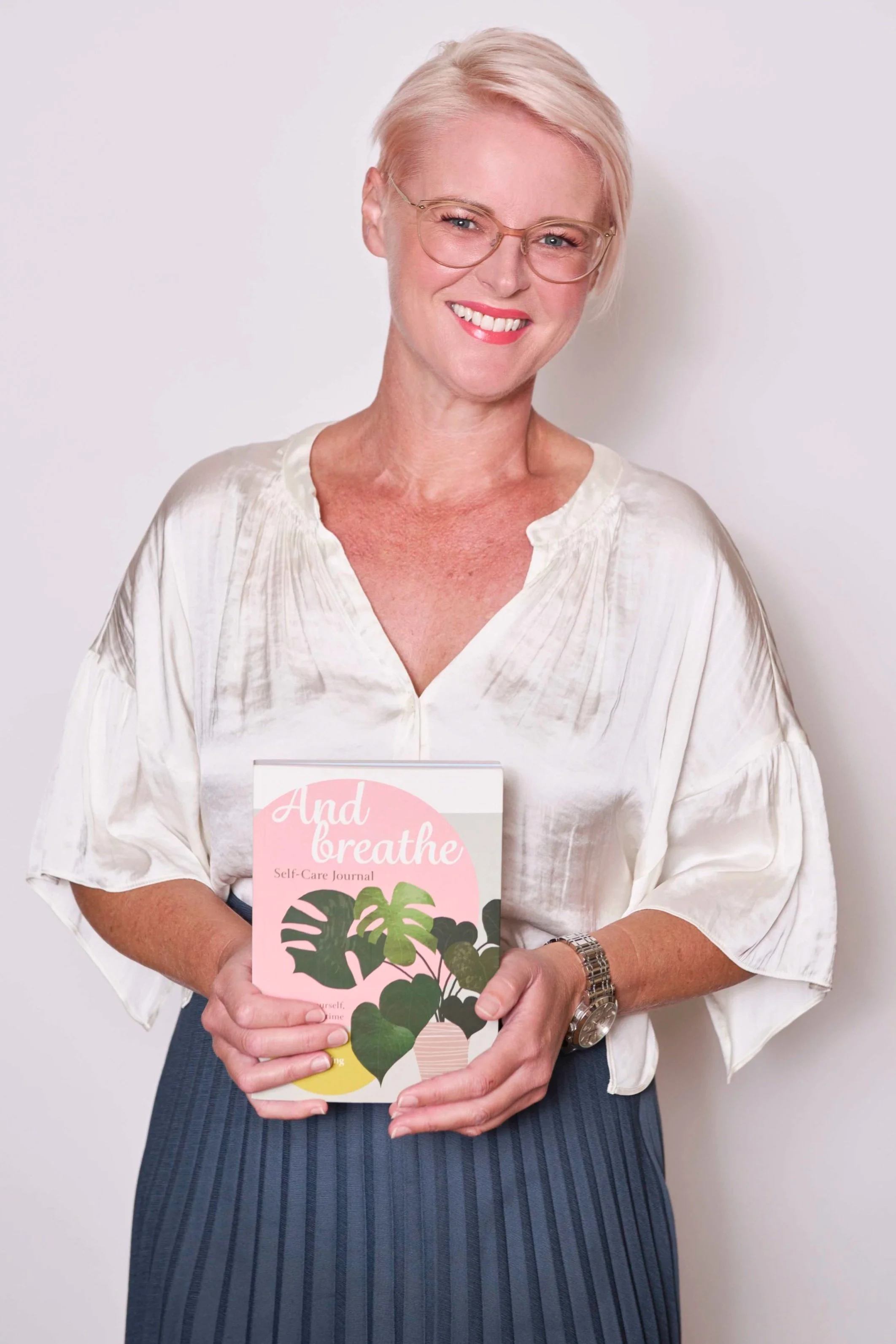 A smiling woman with short blonde hair and glasses holding a self-care journal titled 'Add to Breathe' with tropical leaf illustrations, standing against a plain white background.