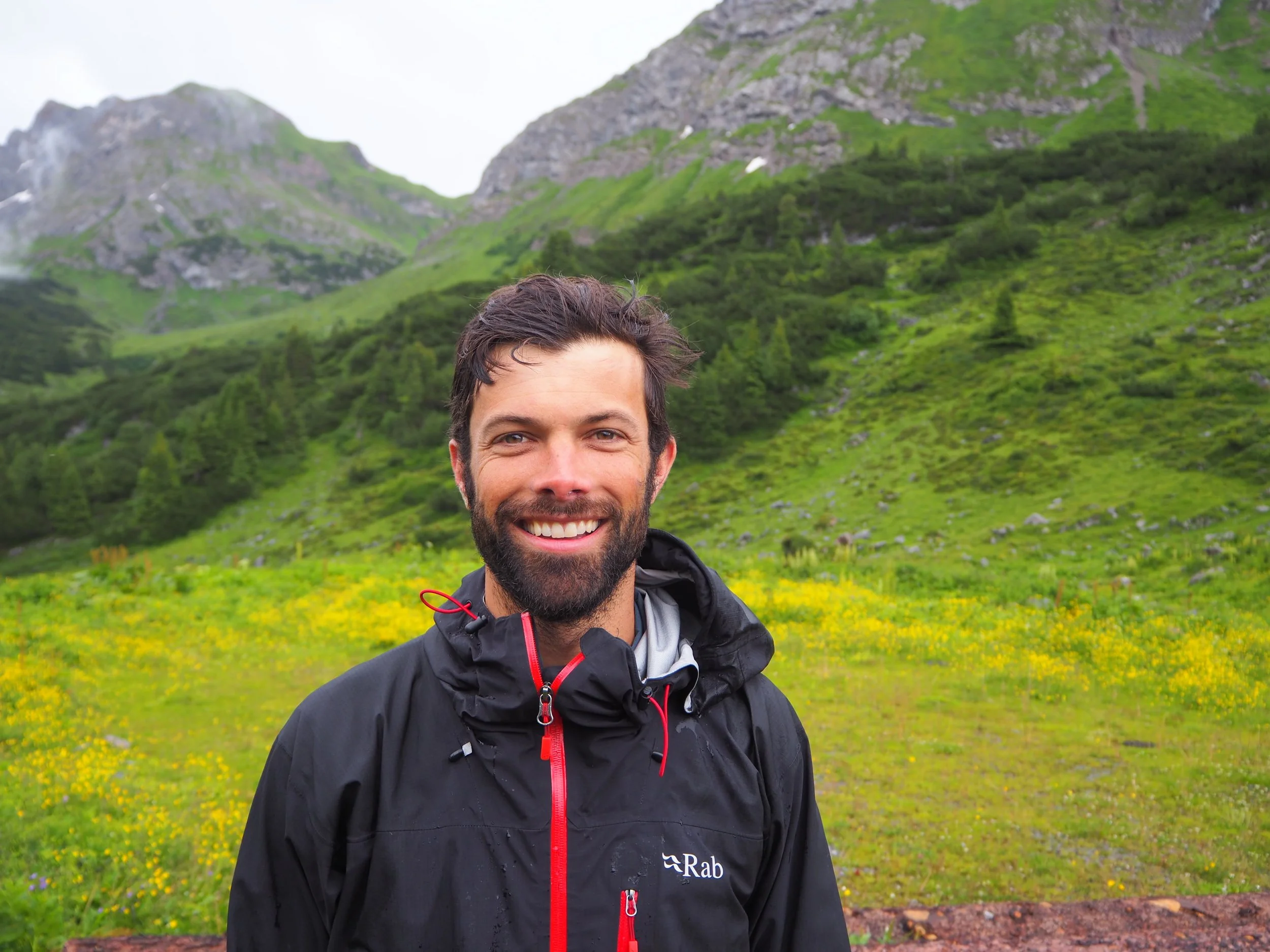 A man with a beard and short dark hair smiling outdoors in front of green mountains and a grassy trail, wearing a black rain jacket.
