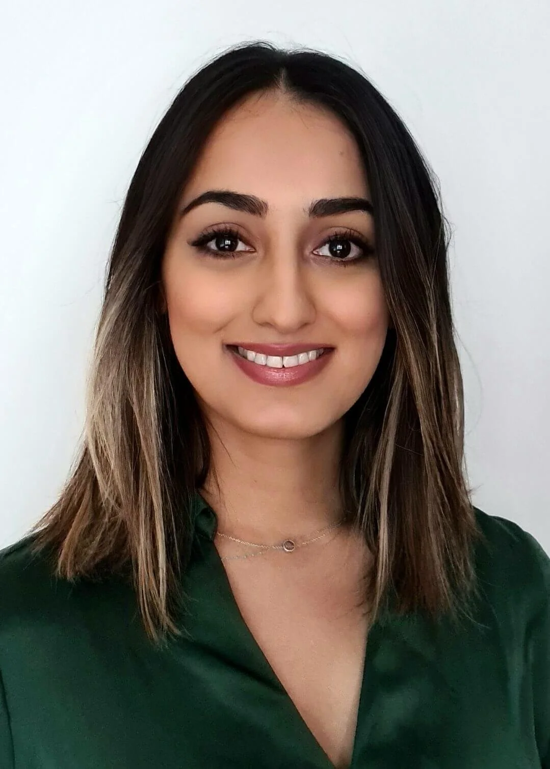 A woman with shoulder-length brown hair, wearing a dark green top, smiling with white teeth, and wearing a delicate necklace, against a plain white background.
