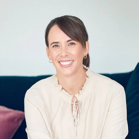 A woman with short dark hair, wearing a white blouse with ruffled details, smiling while sitting on a sofa with a purple pillow and a dark blue throw pillow.