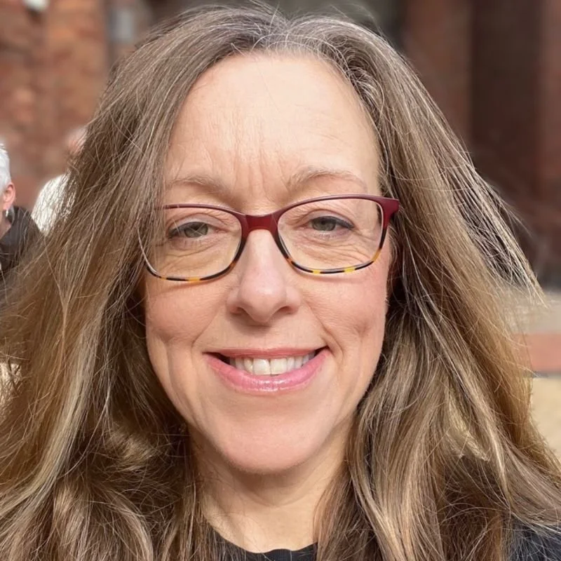 Close-up of a woman with long, wavy brown hair, wearing red glasses, smiling in an outdoor setting.
