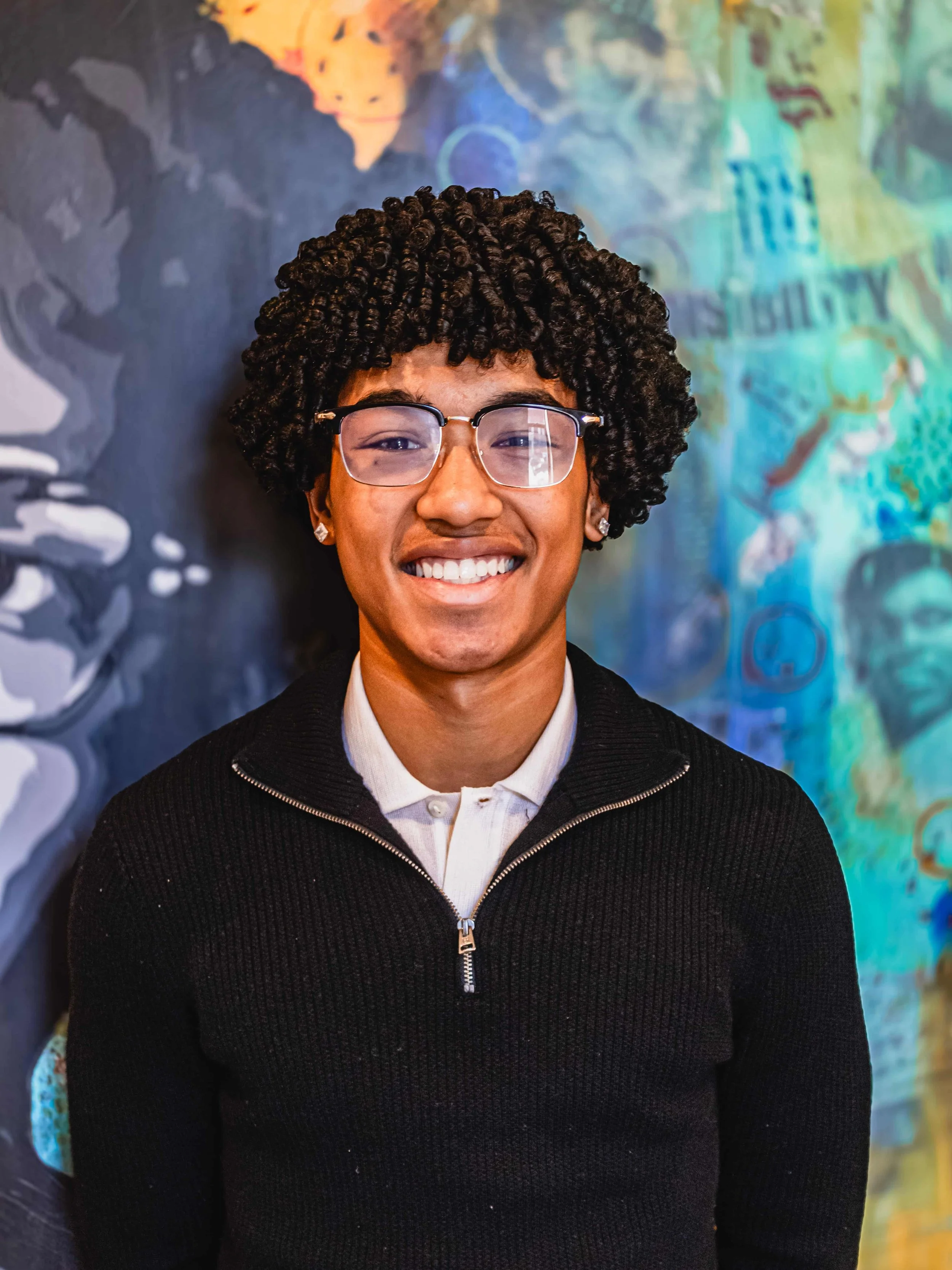 A young person with dark curly hair, glasses, and earrings, smiling and wearing a black zip-up sweater over a white collared shirt, standing in front of a colorful abstract background.