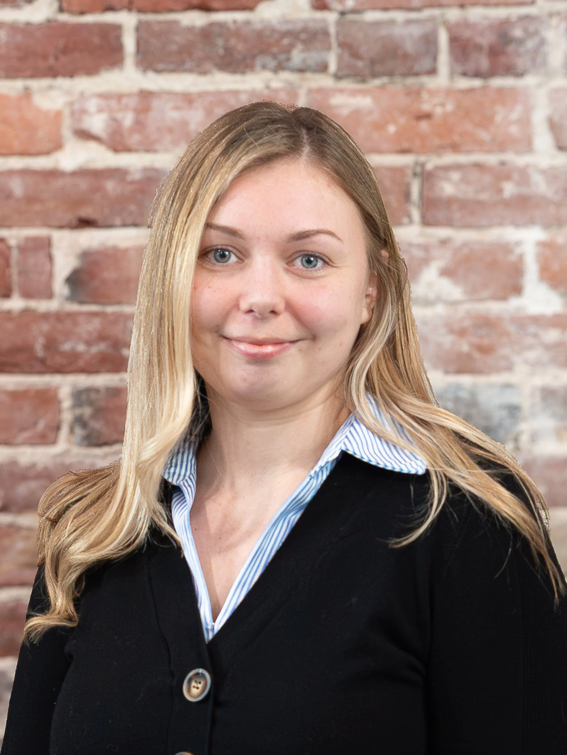 Close-up of a young woman with long blonde hair, blue eyes, smiling, standing in front of a red brick wall, wearing a black coat with a blue striped shirt underneath.