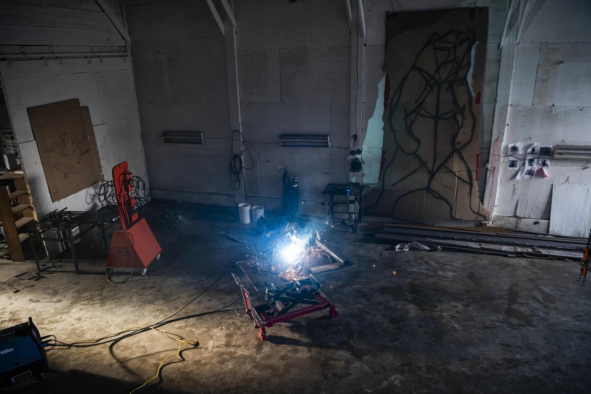 An industrial space with a welding machine sparking, with tools, buckets, and framed artwork on the walls.