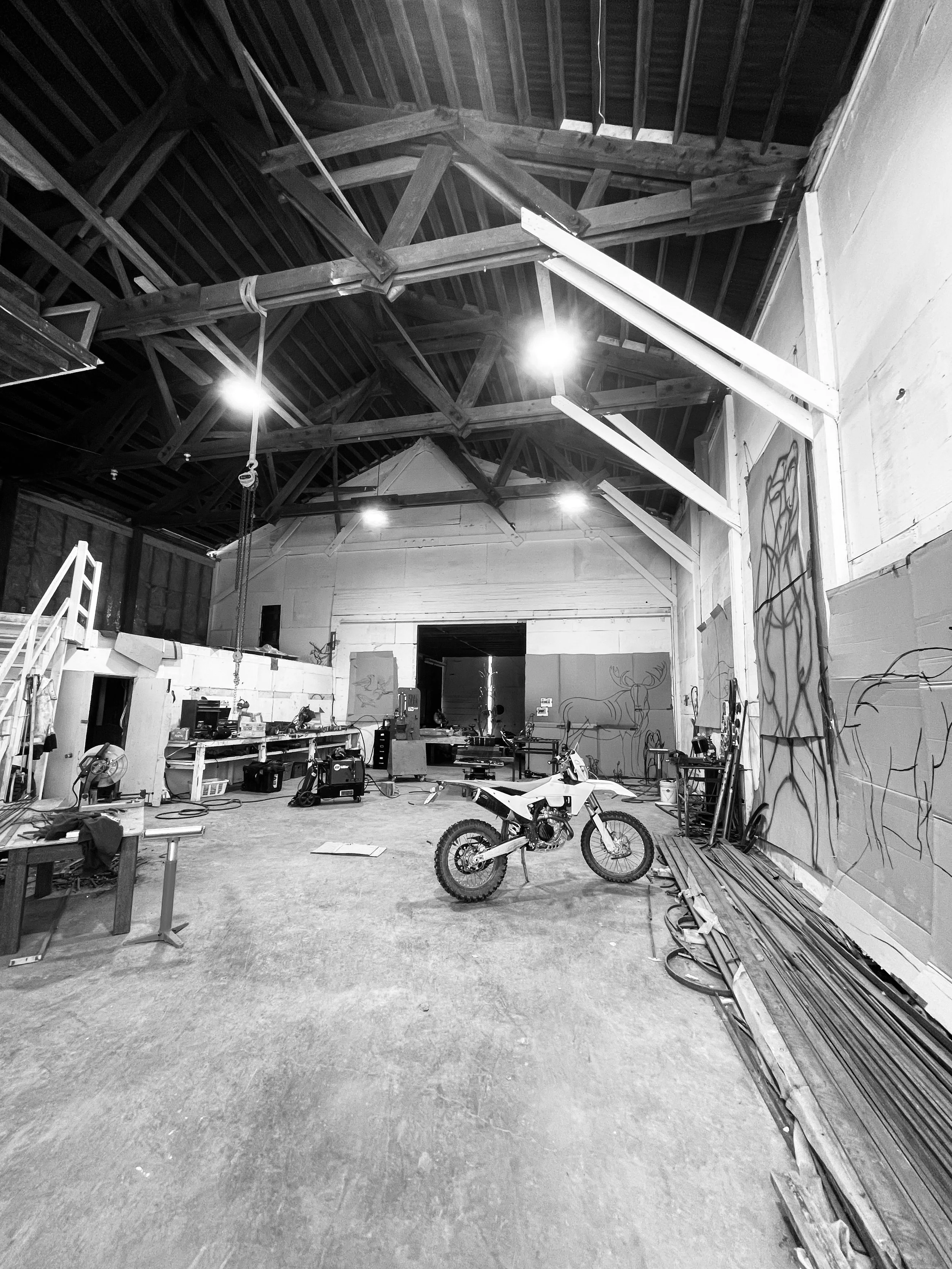 Interior of a workshop or studio with a dirt bike in the center, unfinished walls with graffiti art, workbenches, and construction materials scattered around, wooden beams supporting the high ceiling, and bright overhead lighting.
