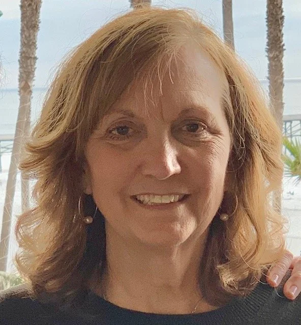 Smiling woman with shoulder-length red hair and earrings, outdoors with palm trees in background.