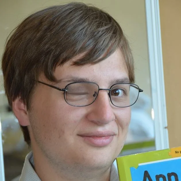 Man with brown hair and glasses winking while holding a colorful book.