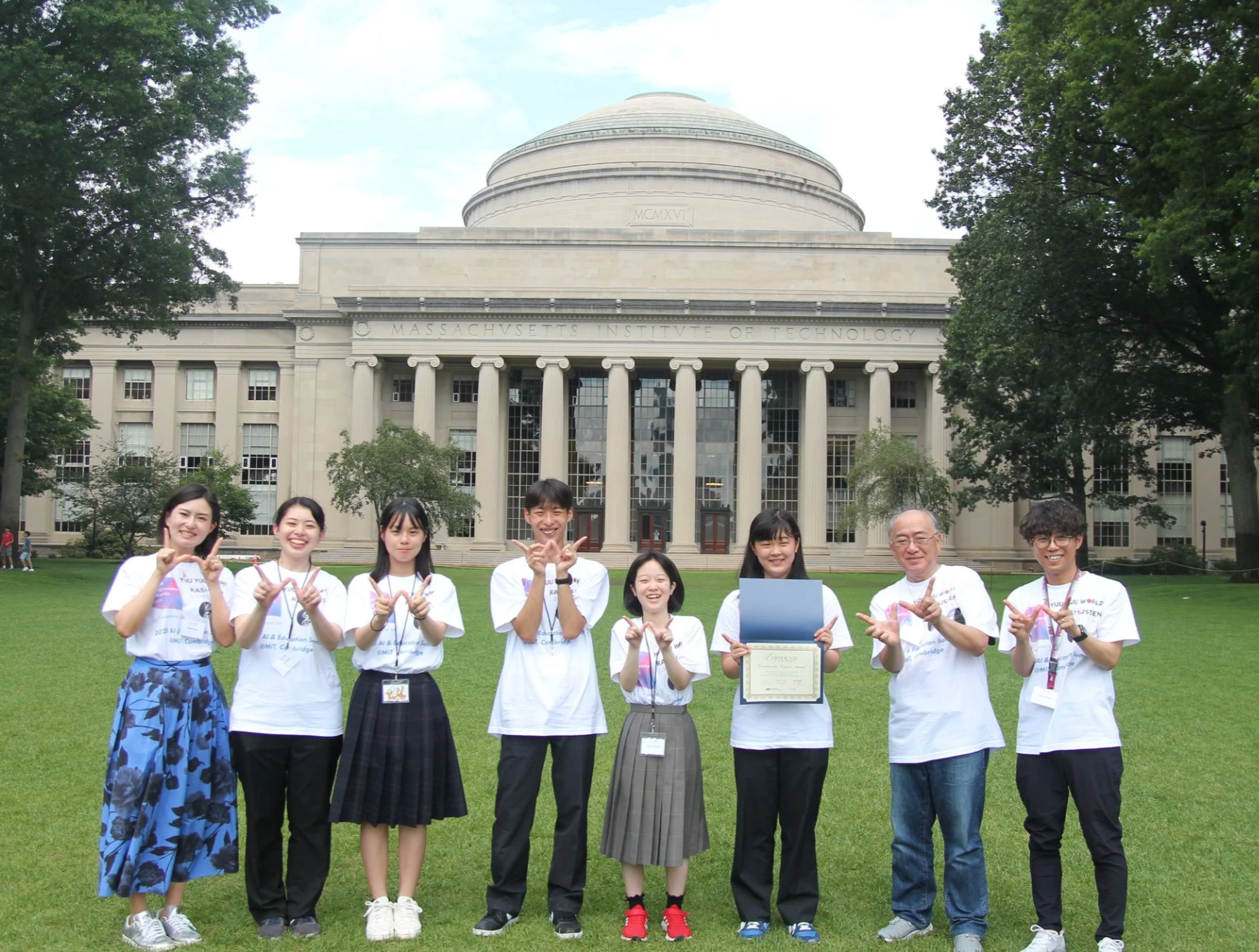 Group of smiling young people standing on grass in front of the Massachusetts Institute of Technology (MIT) building, posing for a photo during a celebration or event, some holding certificates.