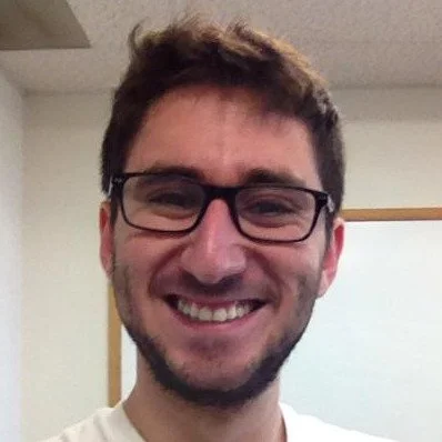 Man with brown hair, glasses, and a beard smiling in an indoor setting.