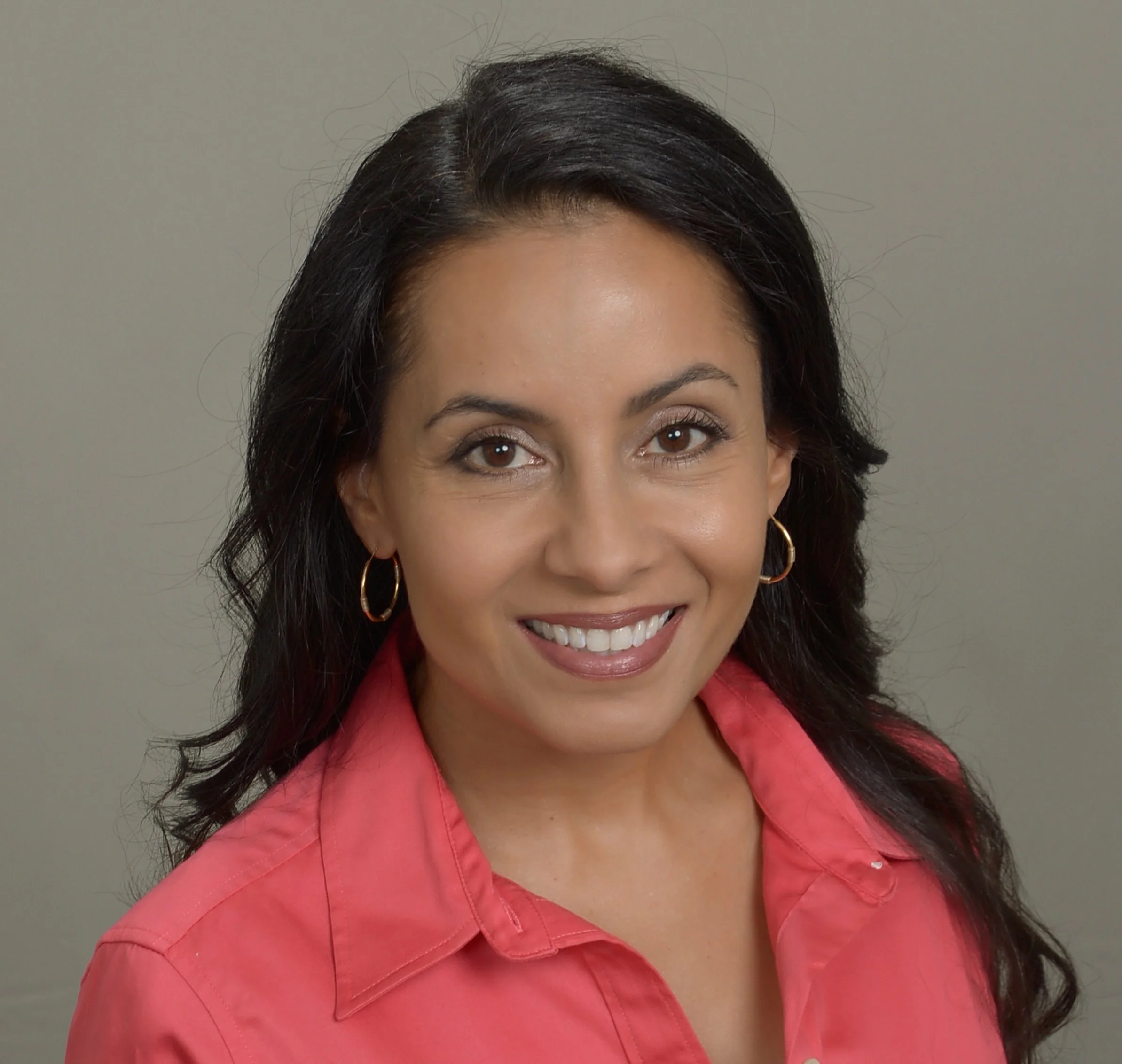 Woman with dark hair, brown eyes, and hoop earrings, wearing a pink blazer, smiling in front of a gray background.