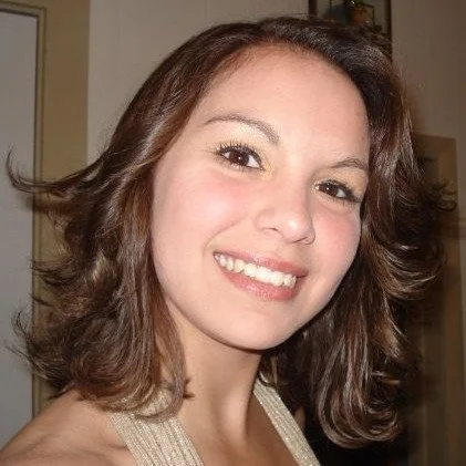 A woman with shoulder-length, wavy brown hair smiling at the camera in an indoor setting.