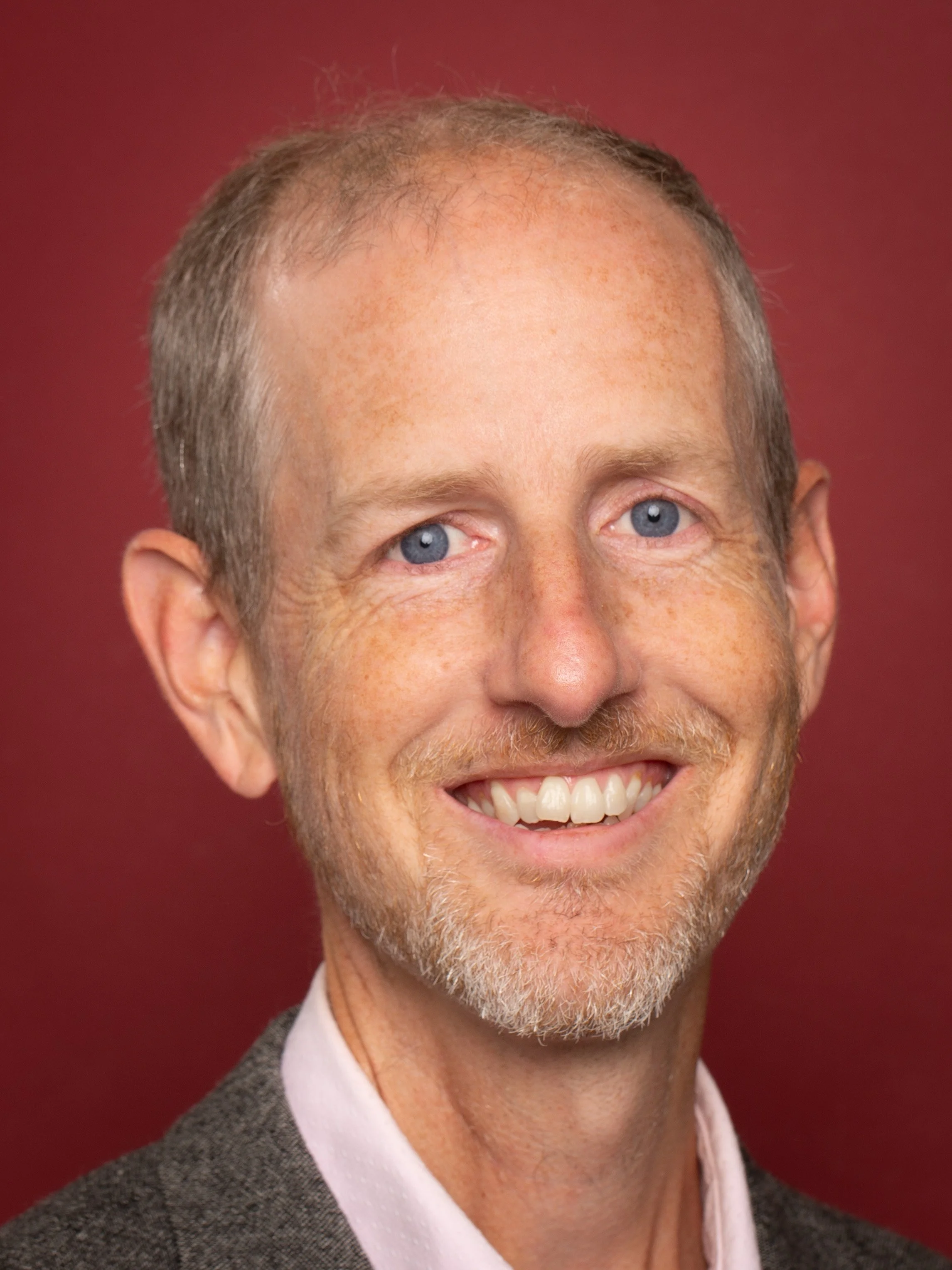 Portrait of a man with light complexion, short brown hair and blue eyes, wearing a blazer and white shirt, against a red background.