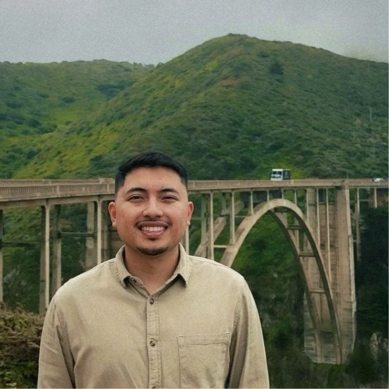 Man standing outdoors in front of a large green hill and an arched bridge, smiling at the camera.