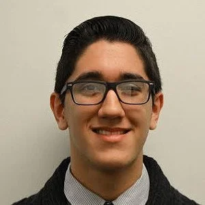 A man with dark hair, glasses, and a light-colored shirt, smiling against a plain light background.
