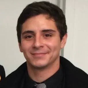 Portrait of a man with dark hair wearing a black shirt and gray tie, smiling softly.