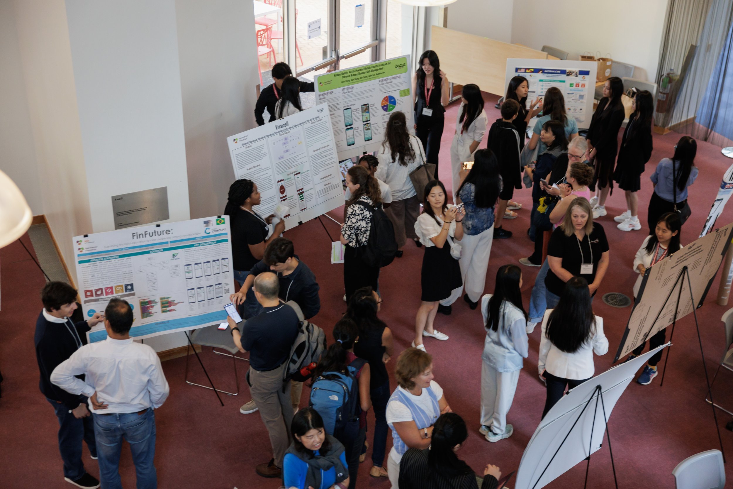 A science conference or academic poster presentation event with multiple research posters displayed on boards, surrounded by attendees, including students and adults, engaging in discussions and viewing research work.