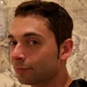 Close-up of a man with dark hair and light skin, smiling slightly, in front of a granite or marble background.