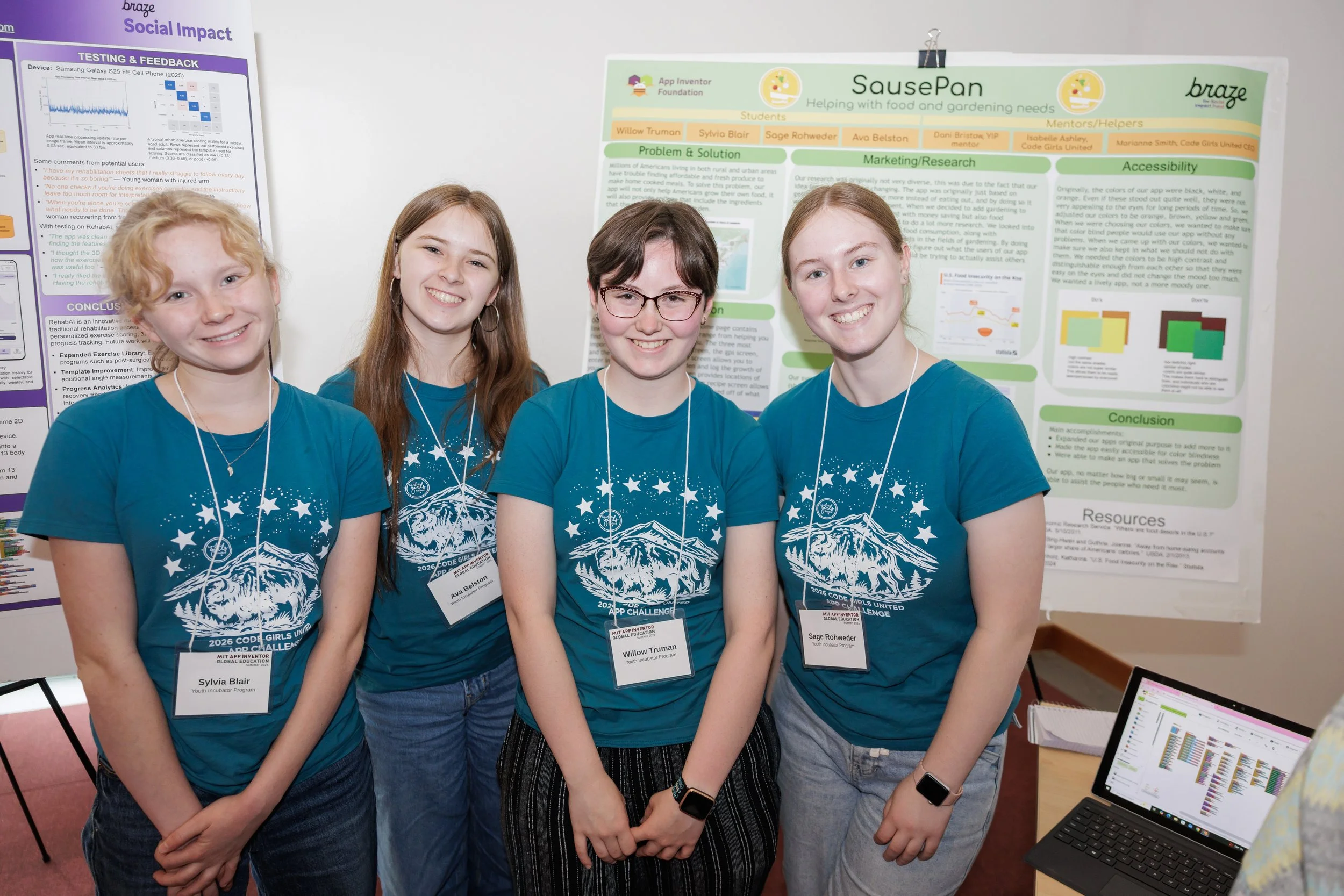 Four girls wearing matching blue t-shirts with a mountain and stars design, standing in front of research posters at a conference, smiling at the camera.
