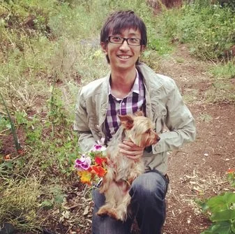 Person holding a small dog and a bouquet of flowers outdoors in a wooded area.