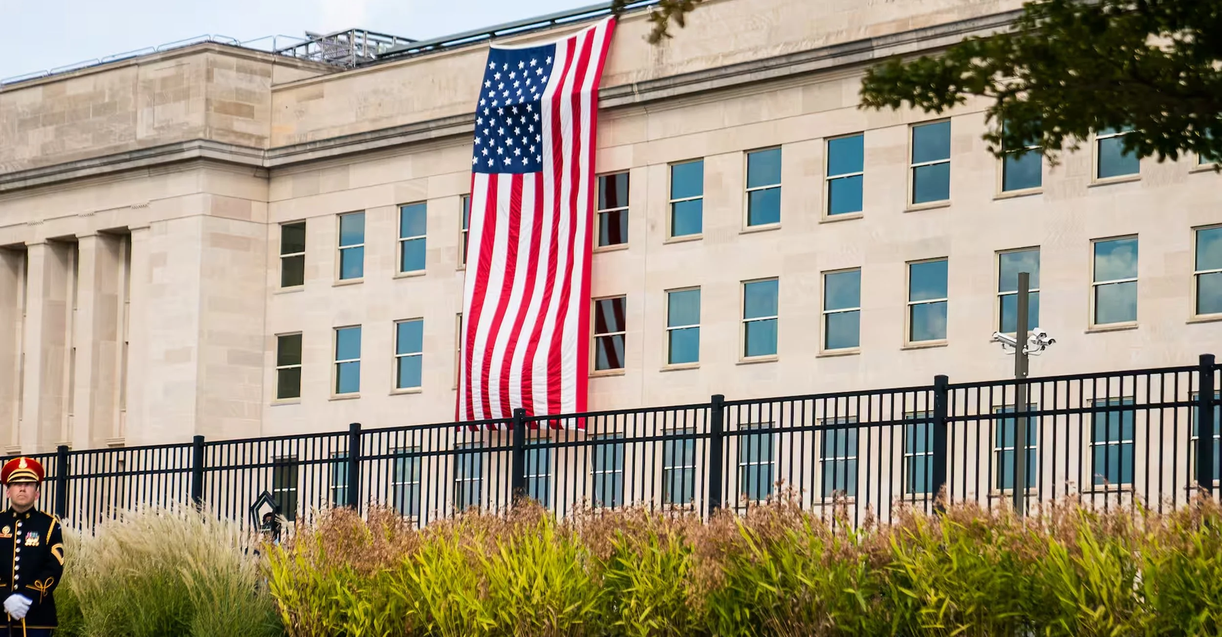 American flag hanging on building with multiple windows, black fence in front, and flagpole with security camera in front, bordering garden with tall grasses and trees.