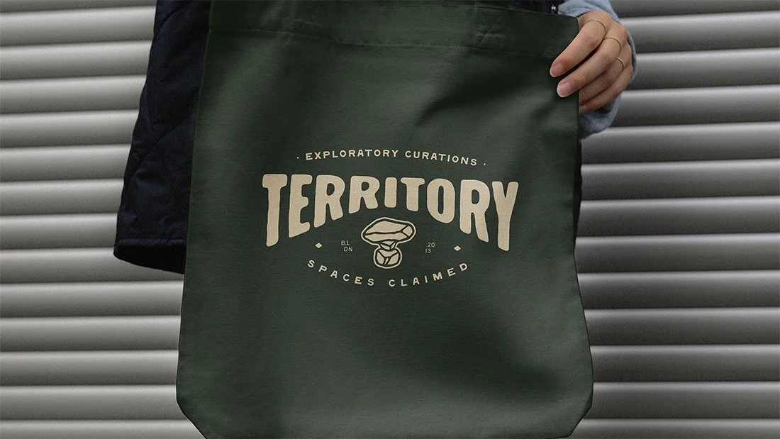 Person holding a dark green tote bag with the logo design for Territory furniture on it, against a background of metal shutters.