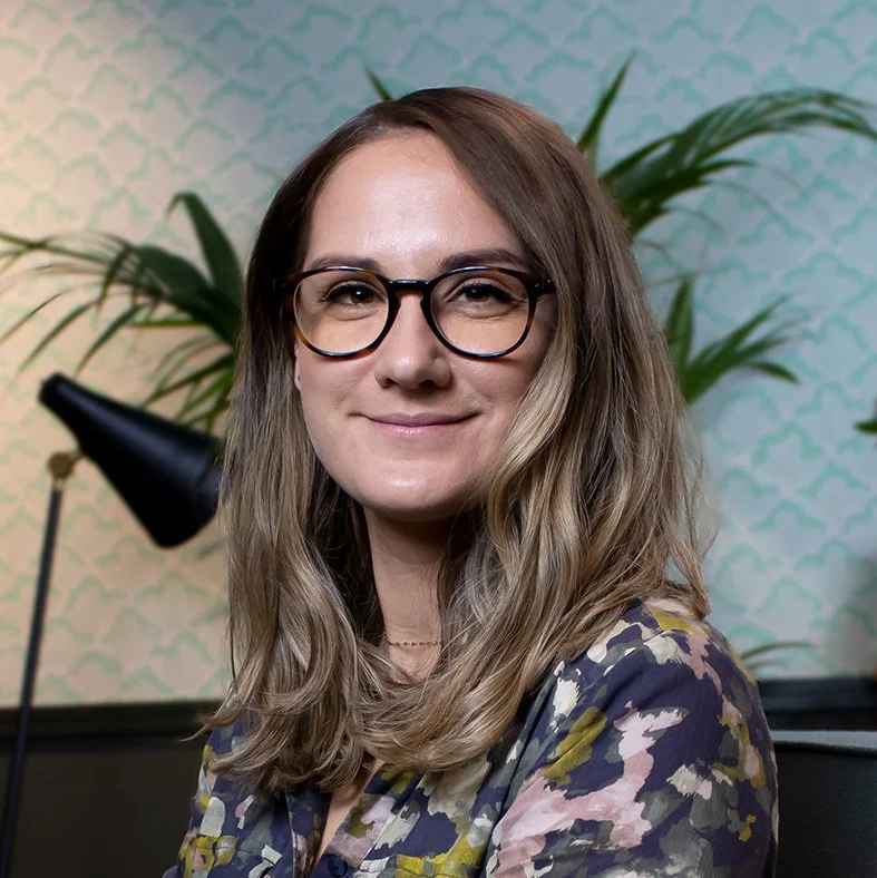 A woman with glasses and long hair, smiling, seated in a room with a patterned wall and plants in the background.