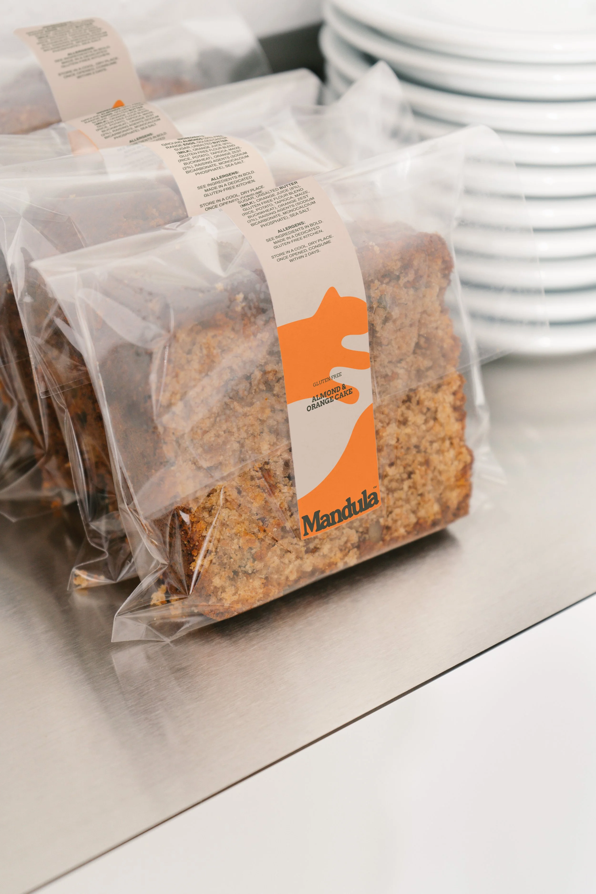 A clear plastic bag with a label that indicates it contains gluten-free almond and orange cake, placed on a metallic surface, with a stack of white plates in the background.