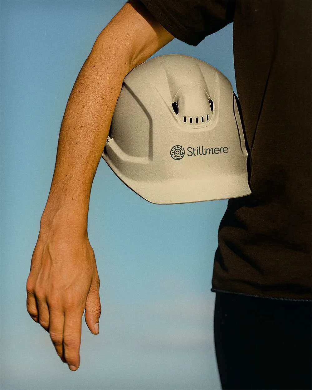 A person holding a hard hat in their arm with the Stillmere logo branded on the front. The person is wearing a black shirt and is standing against a blue background.