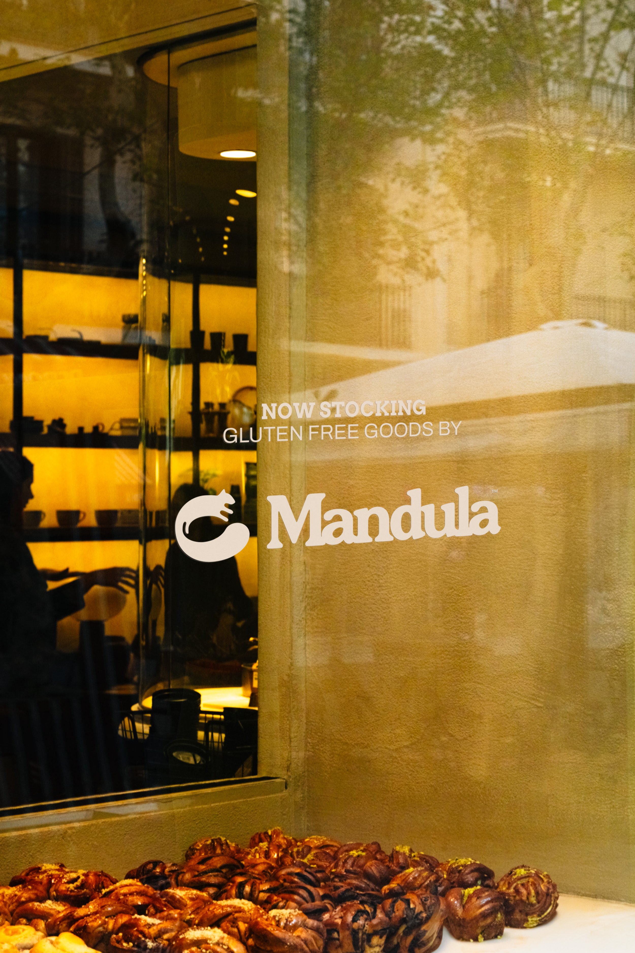 Display of chocolate-covered pastries in a bakery window, with a sign that reads 'Now Stocking Gluten Free Goods by Mandula' and reflections of the bakery interior and street scene.