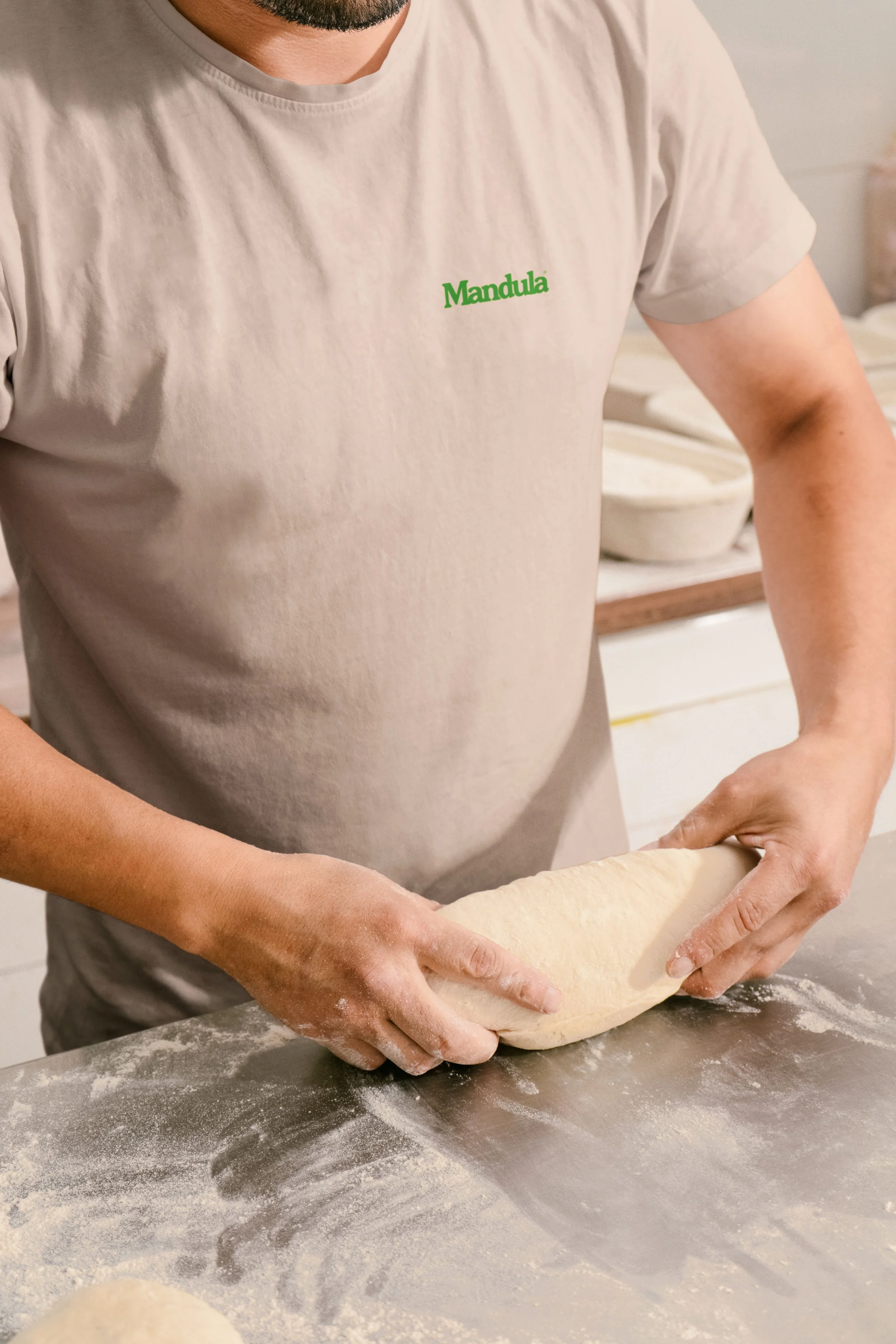 Person kneading dough on a floured surface, wearing a light-colored t-shirt with the word 'Mandula' in green. Hands are shaping the dough, with flour scattered around.