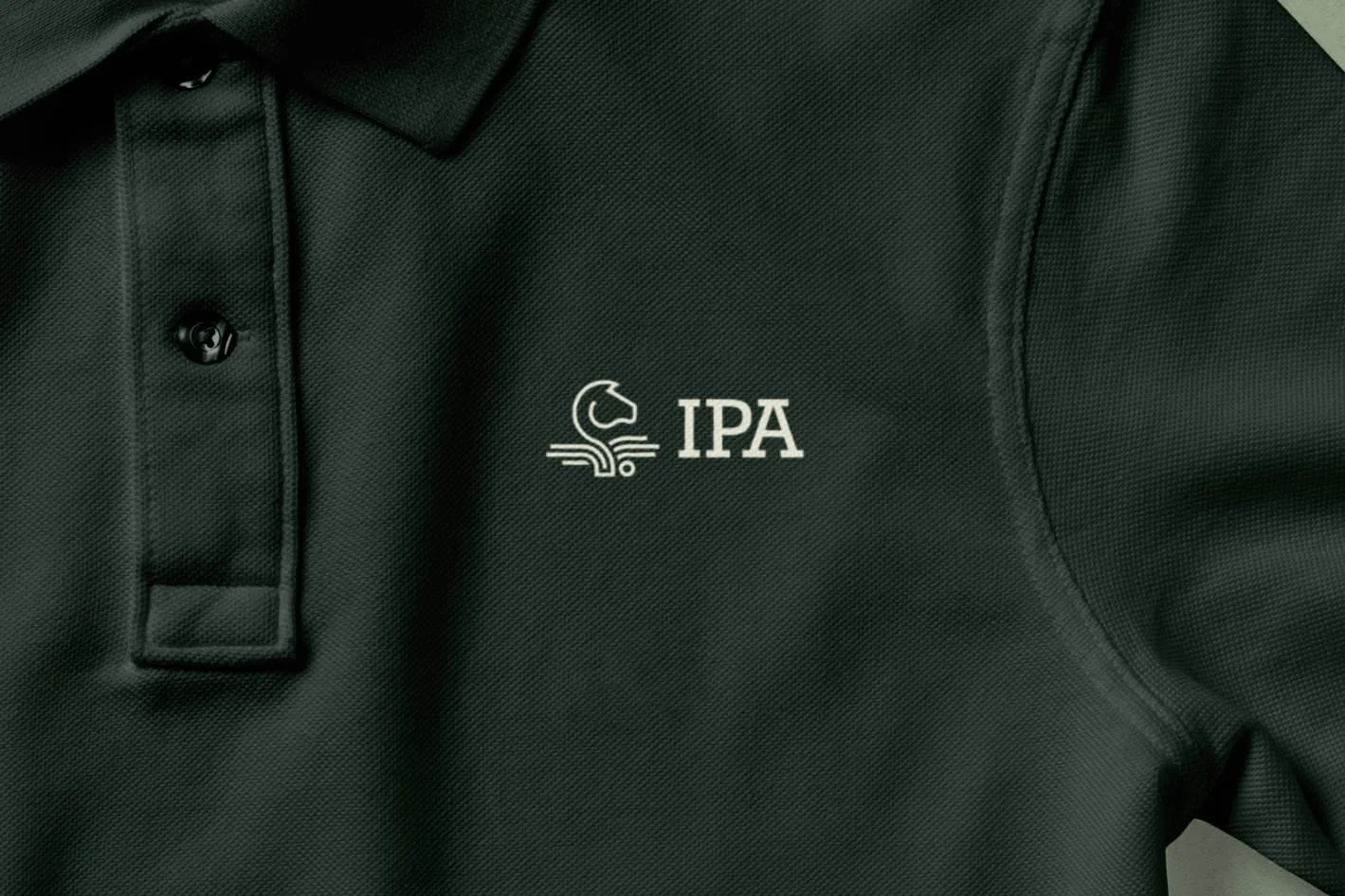 Close-up of a dark green polo shirt with an IPA logo on the left chest, featuring a horse head and stylised book.