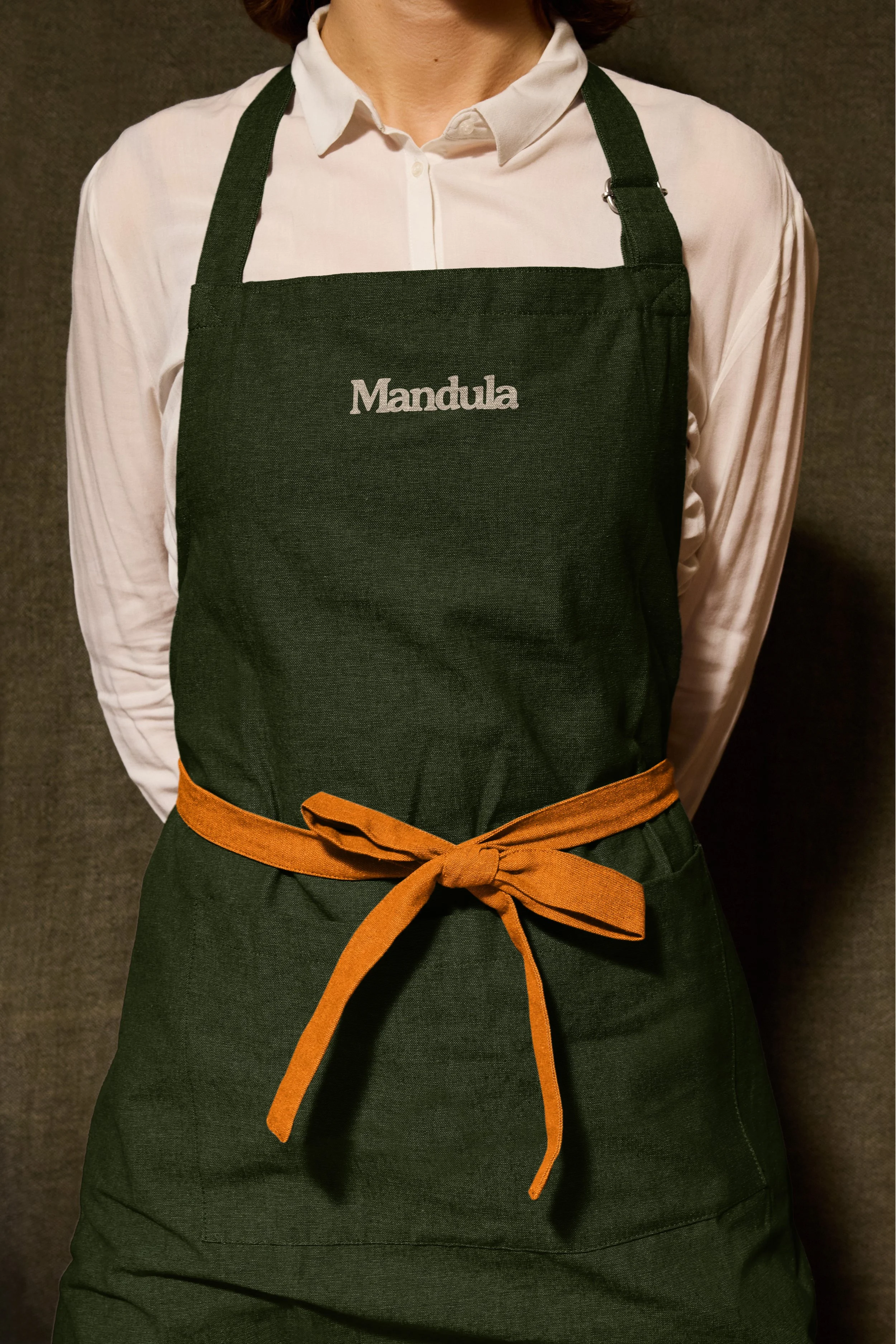 Person wearing a white shirt with a black apron that has the word 'Mandula' embroidered on it, and a brown waist apron tied at the front.