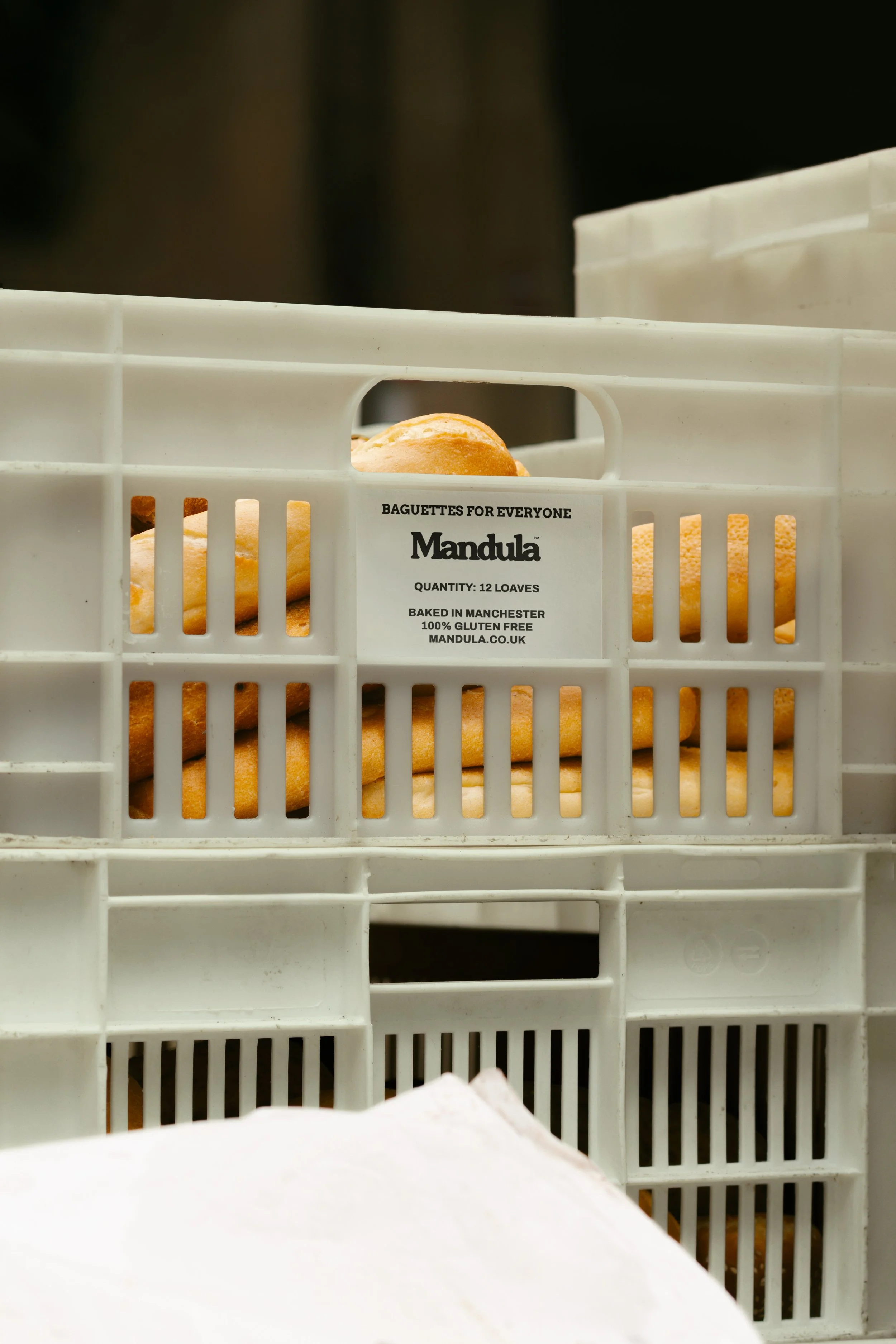 White plastic crate containing baked baguettes with a label indicating they are gluten free, baked in Manchester, quantity of 12 loaves.