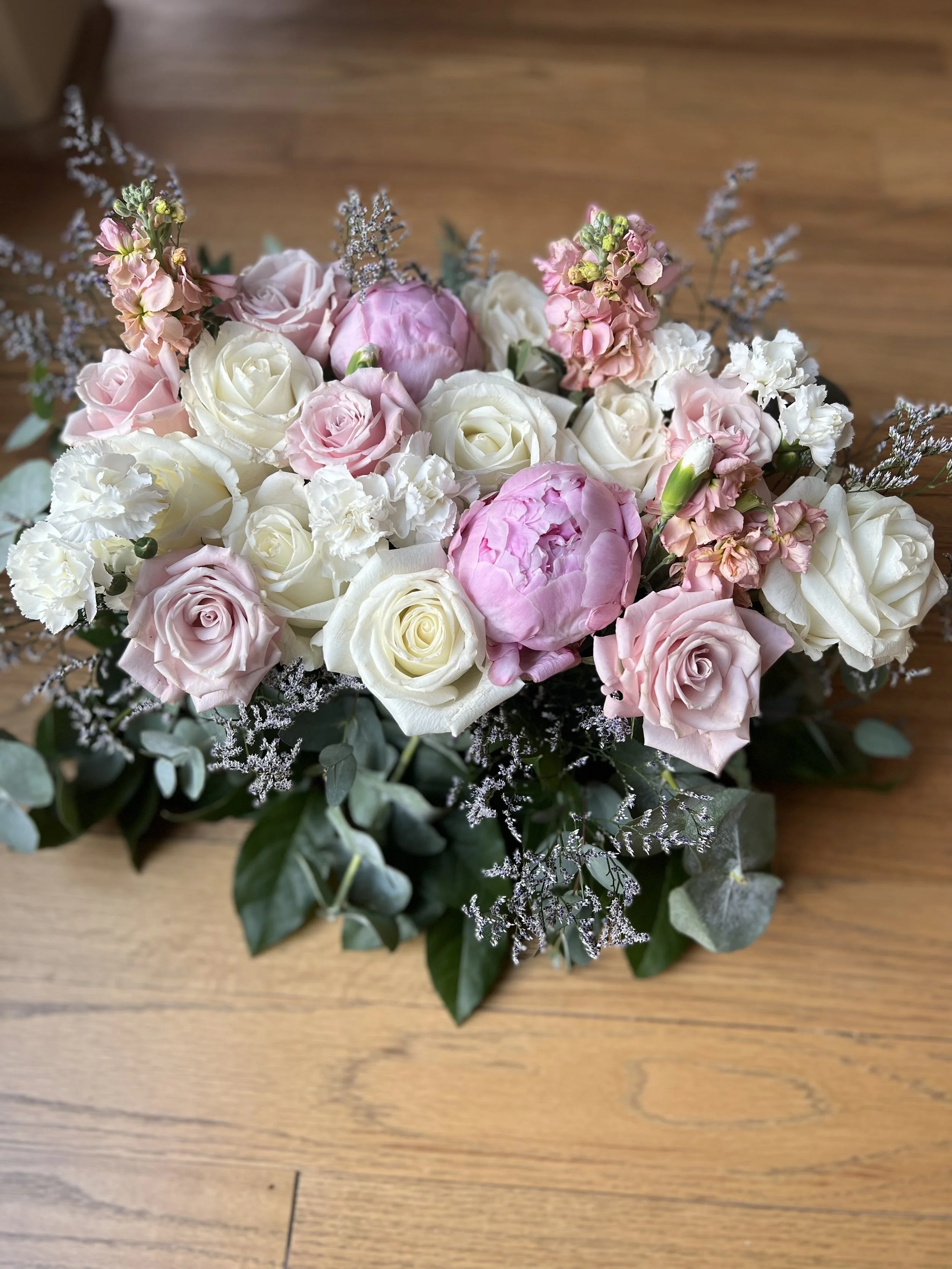 Bouquet of white and pink roses, peonies, and carnations with greenery on a wooden floor.