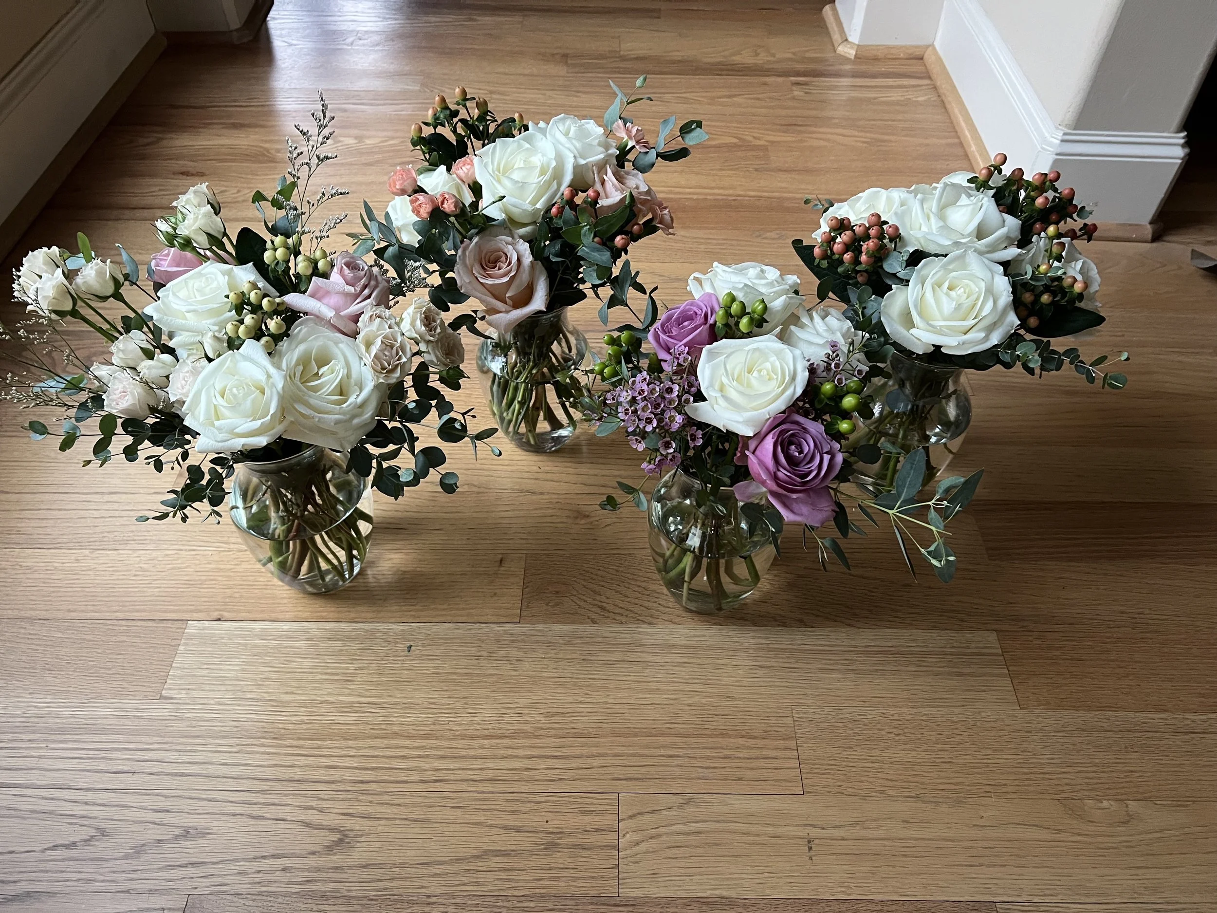 Four glass vases on a wooden floor, each holding a bouquet of flowers including white roses, purple roses, small berries, and green foliage.