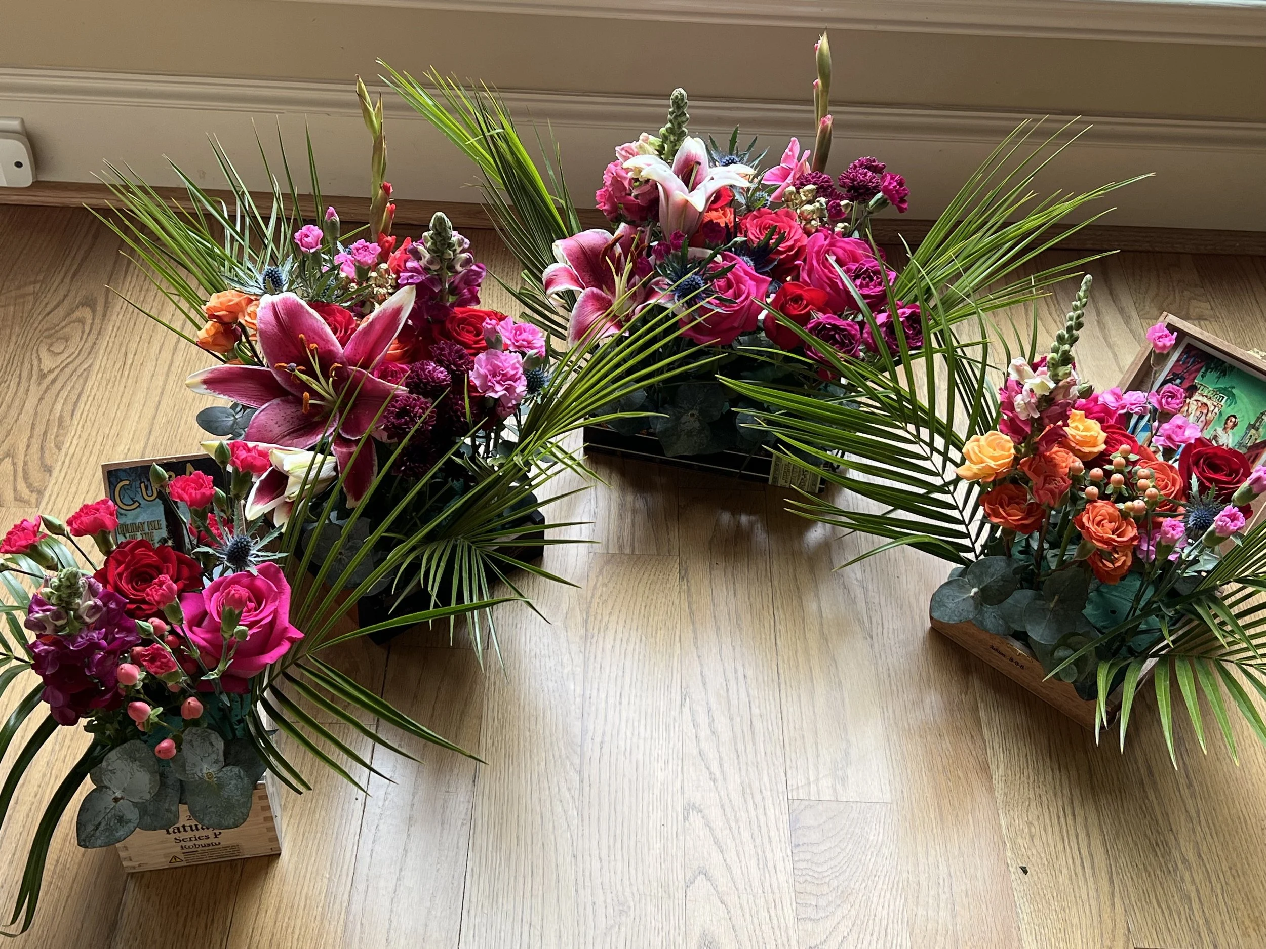 Floral arrangements with pink lilies, roses, snapdragons, and palm leaves on wooden floor.