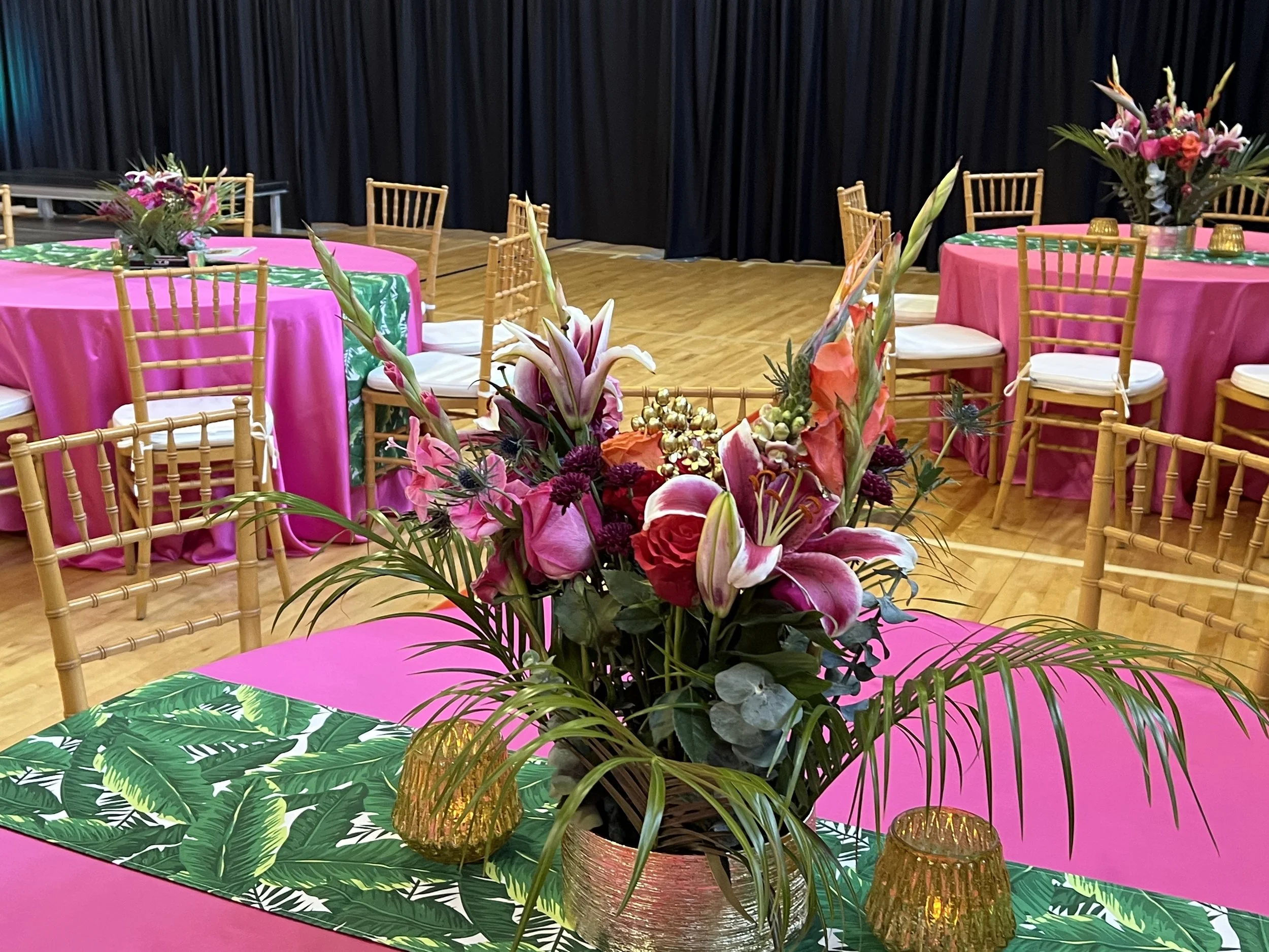 Event hall with tables covered in pink tablecloths, green leaf-patterned runners, and floral centerpieces. Wooden chairs surround the tables.