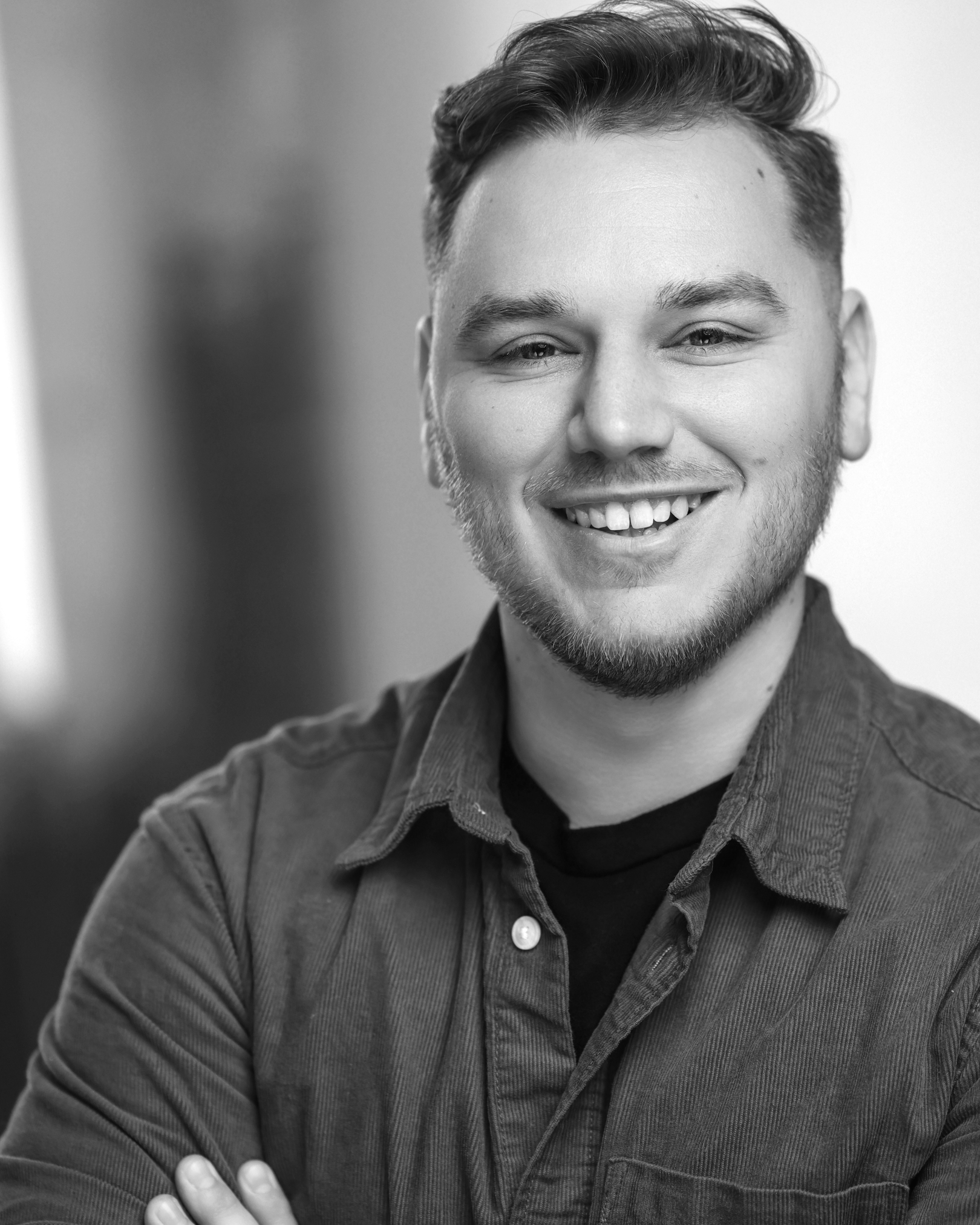 Black and white portrait of a smiling young man with a beard and wavy hair, wearing a T-shirt, against a dark background.
