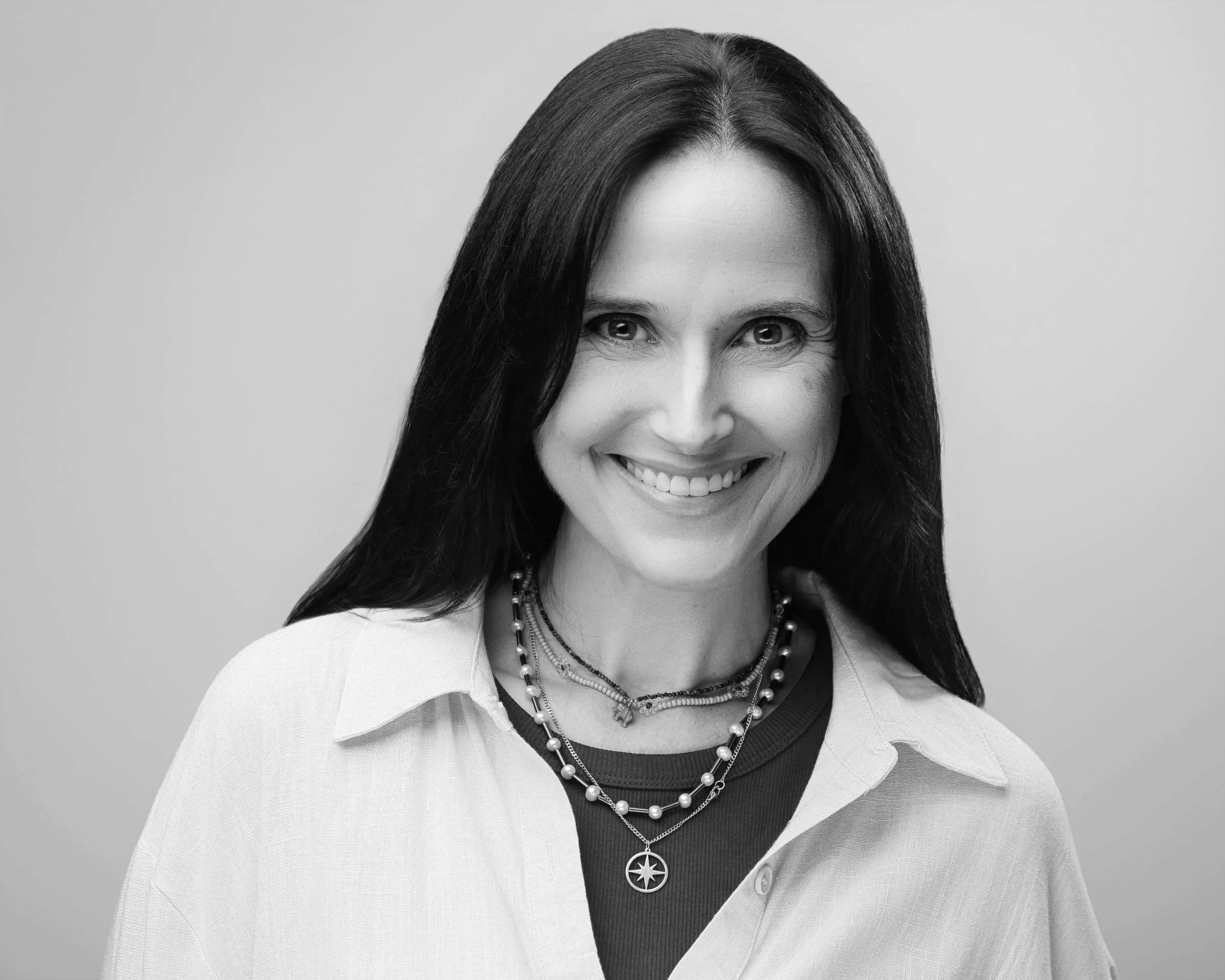Black and white portrait of a smiling woman with dark, shoulder-length hair, wearing a sleeveless black top, against a plain background.