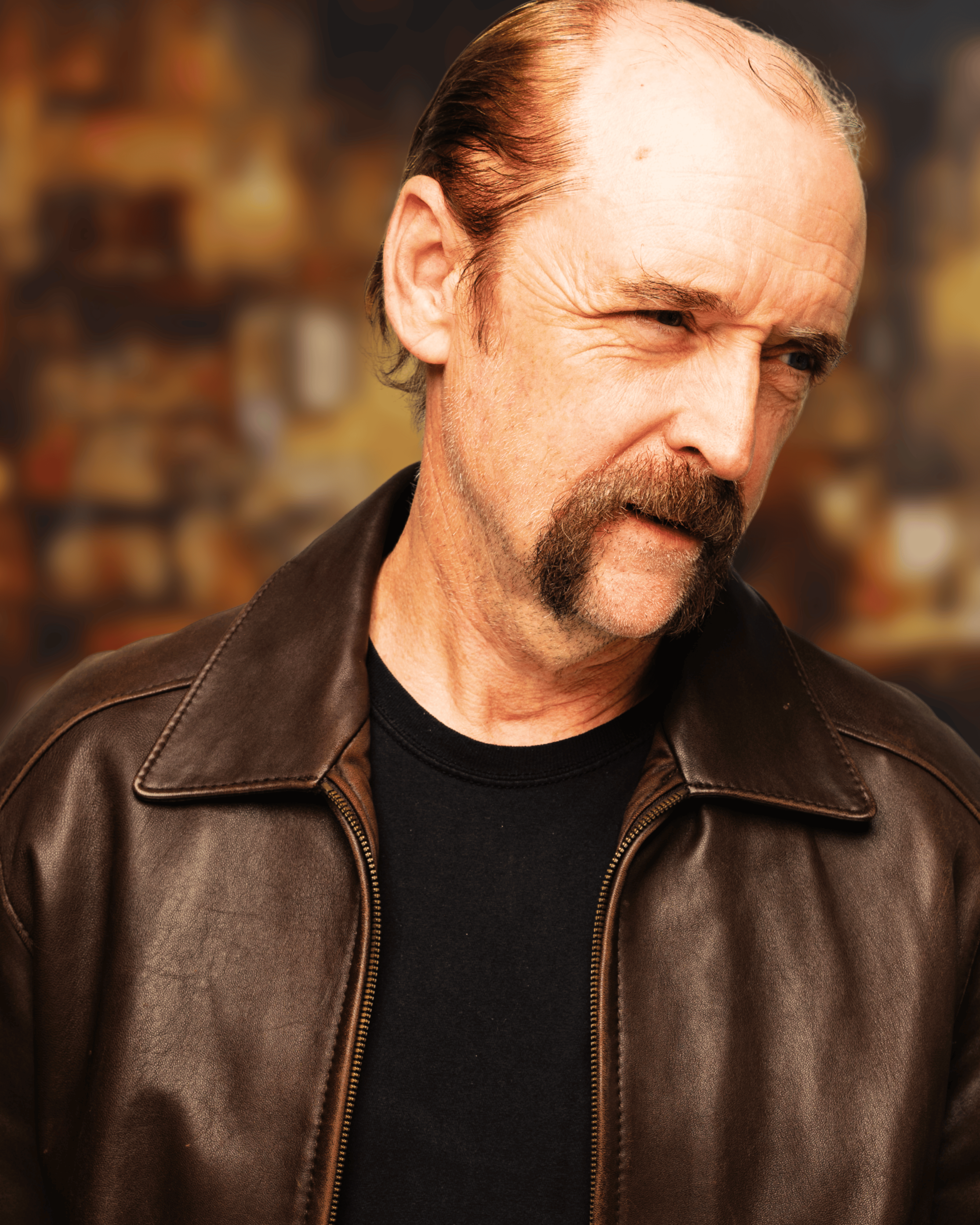 A middle-aged man with a full beard and mustache, wearing a brown leather jacket over a black shirt, looking to his right with a thoughtful expression. The background is blurred and warm-toned.