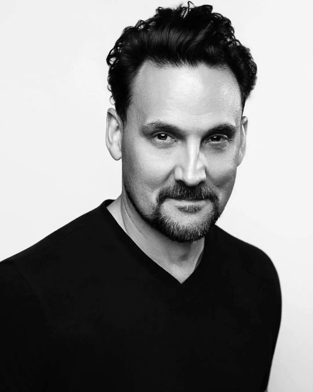 Black and white portrait of a man with short dark hair, styled upward, light eyes, a beard, and a mustache, wearing a textured, dark shirt, against a dark background.