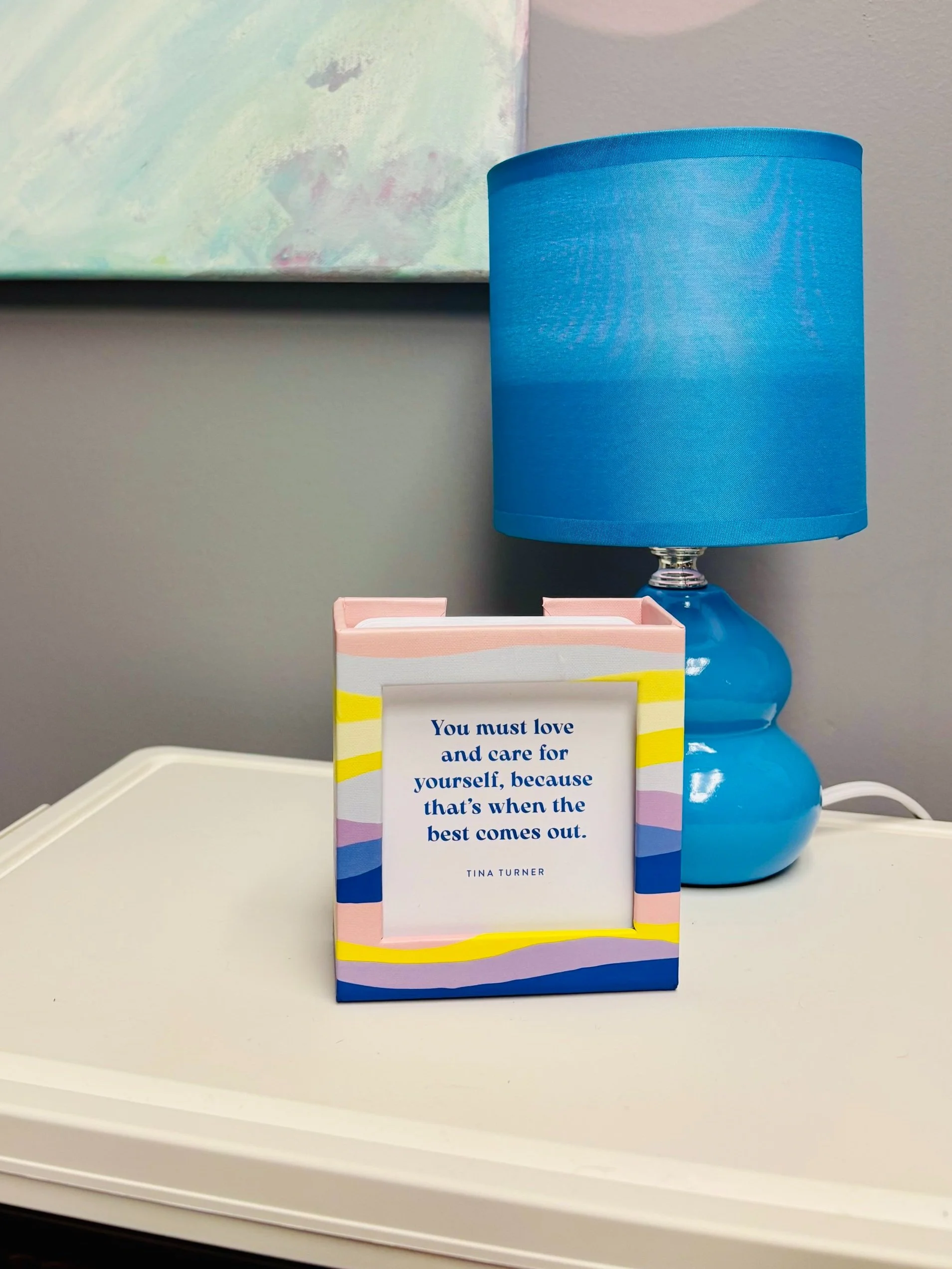 Picture of a small sign that reads "you must love and care for yourself, because that's when the best comes out. -Tina Turner." Behind that a small blue lamp and the bottom of an abstract painting, hung on a gray wall.