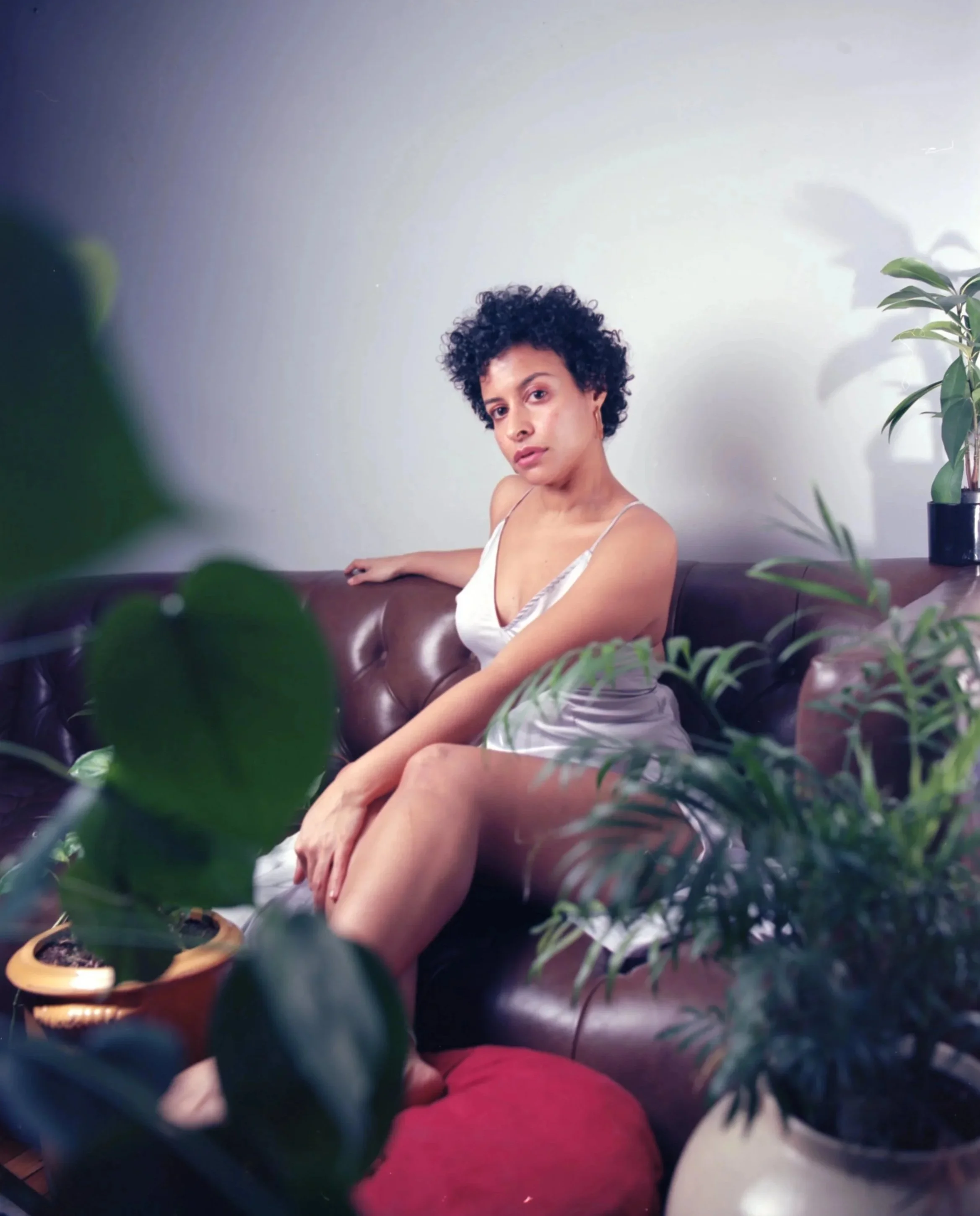 A woman with short curly hair sitting on a brown leather sofa, surrounded by green houseplants, wearing a silver slip dress and looking at the camera.