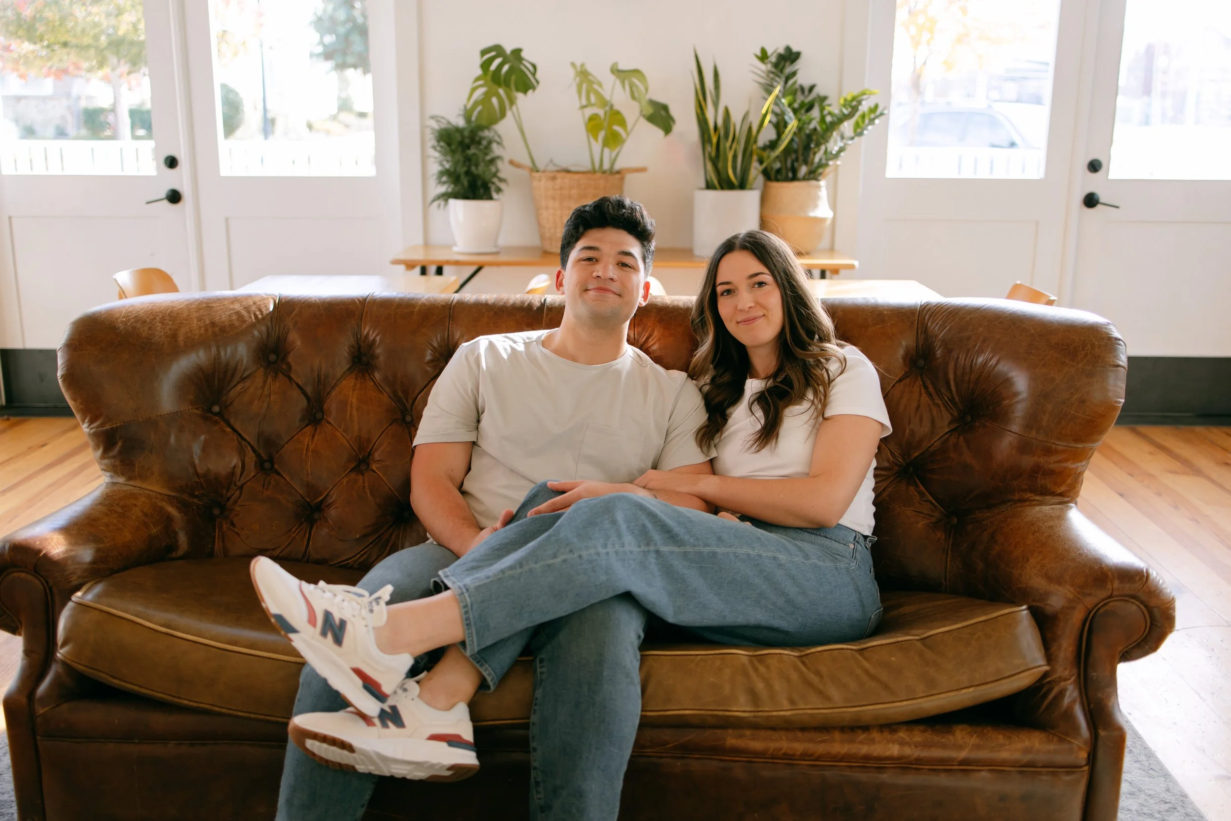 A young man and woman sitting closely together on a brown leather couch in a brightly lit room with large windows and green plants at the back. They are smiling and relaxed.