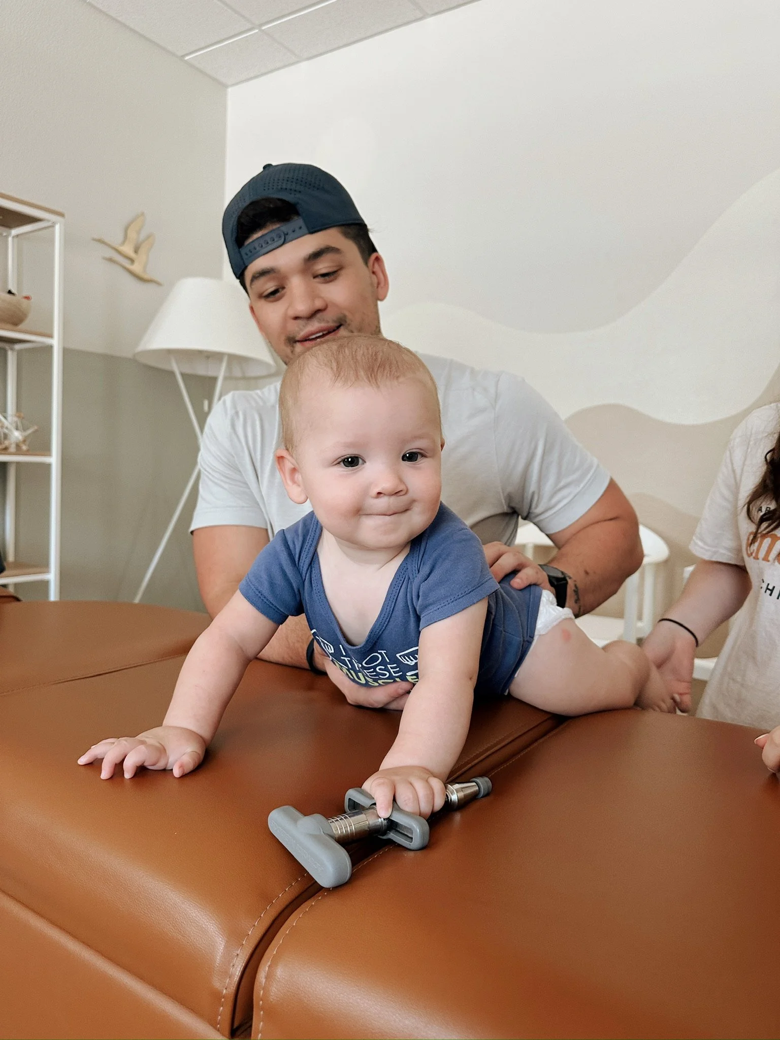Baby lying on a brown surface with a person in a baseball cap.