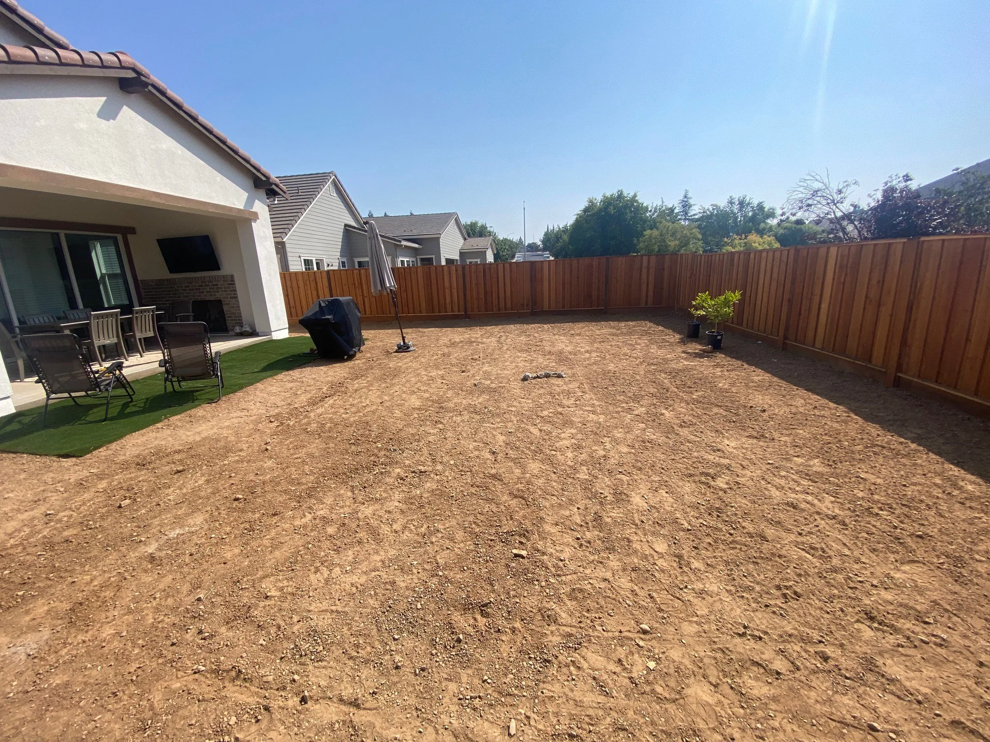 Backyard with dirt ground, a section of artificial grass, patio furniture, a grill, a patio umbrella, and a wooden fence.