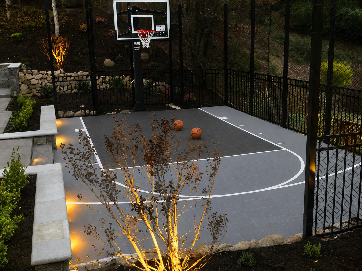 An outdoor basketball court with two basketballs on the gray playing surface, surrounded by a black metal fence, with trees and plants around it, and illuminated by soft lighting at dusk.